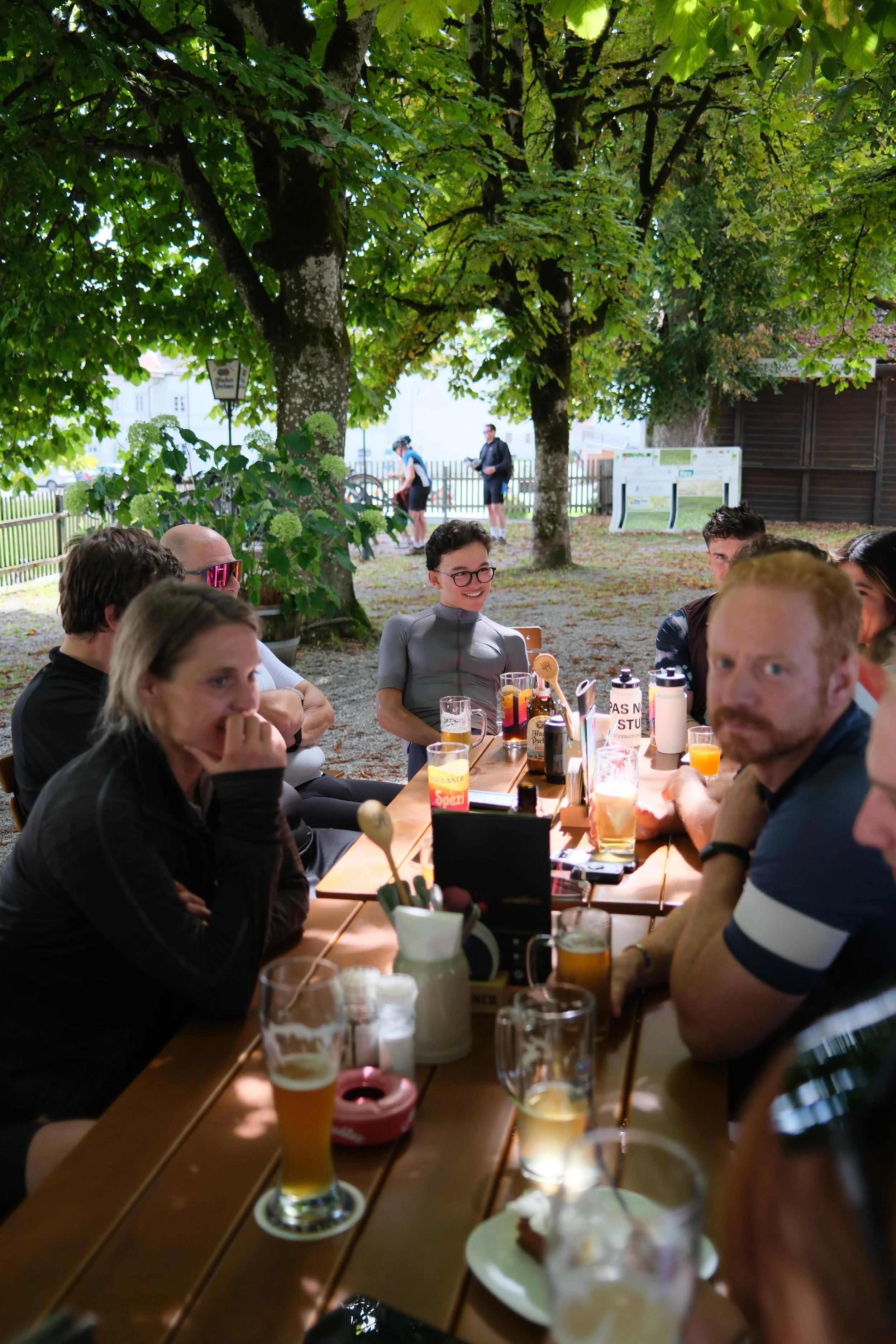 Gruppe von Leuten, die an einem langen Holztisch im Freien sitzen, trinken und sich unterhalten, umgeben von Bäumen und einer Gartenumgebung.