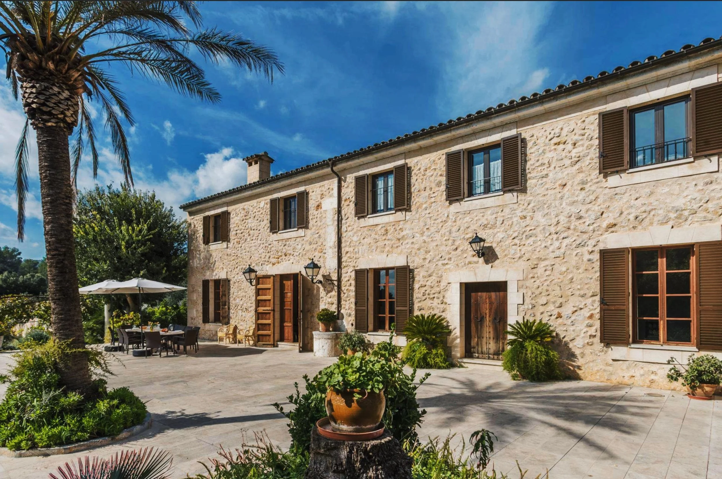 Ein malerisches Haus mit Steinmauern, braunen Fensterläden und Holzportalen. Im Vordergrund ist ein gepflasterter Hof mit Sitzgelegenheiten, einem großen Sonnenschirm und zahlreichen Topfpflanzen, umgeben von Palmen und üppigem Grün. Der Himmel ist blau mit wenigen Wolken.