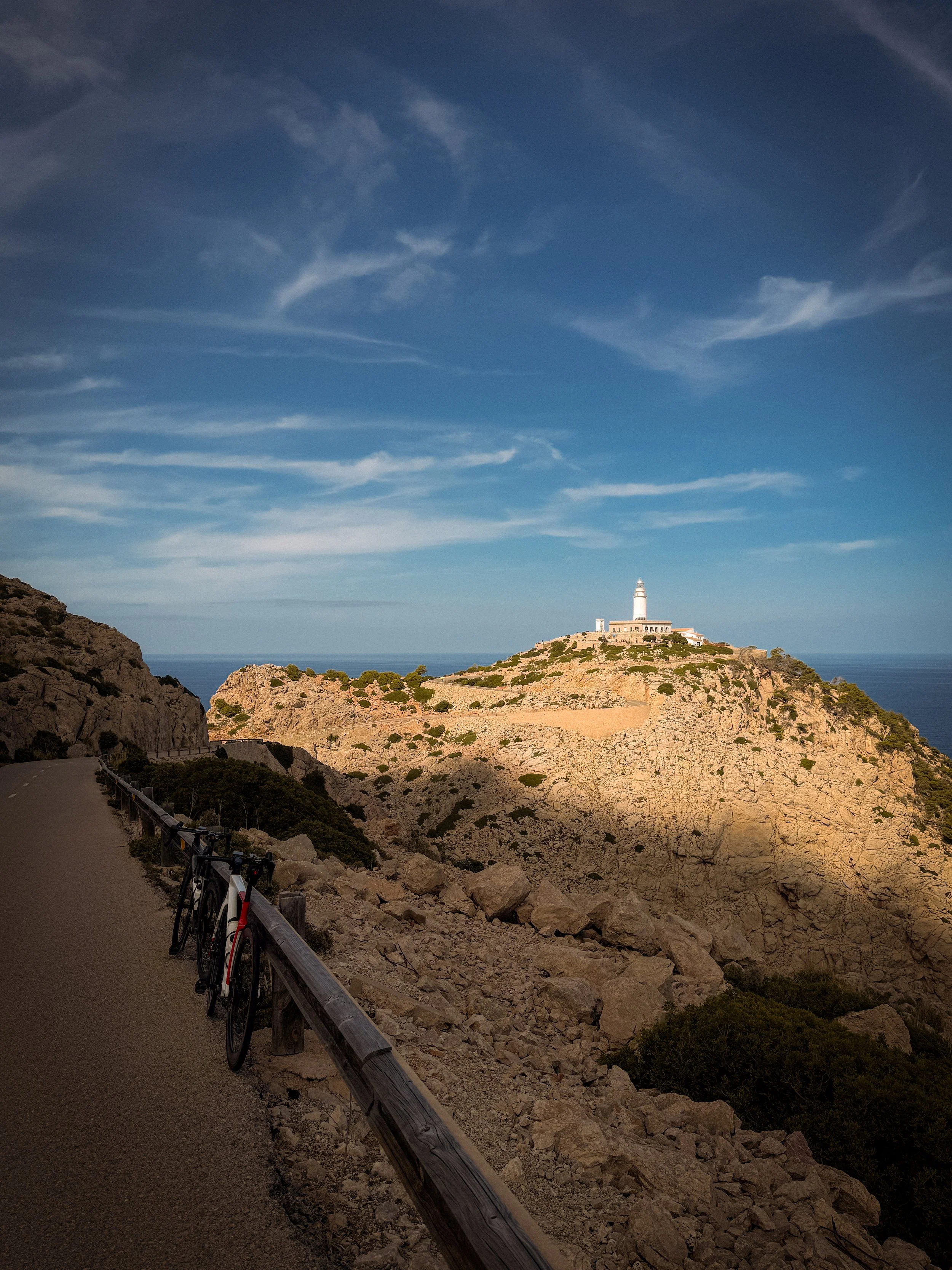 Straße mit Rennrädern am Rand, Blick auf den Leuchtturm von Cap Formantor auf einer Klippe bei schönem Himmel auf Mallorca.