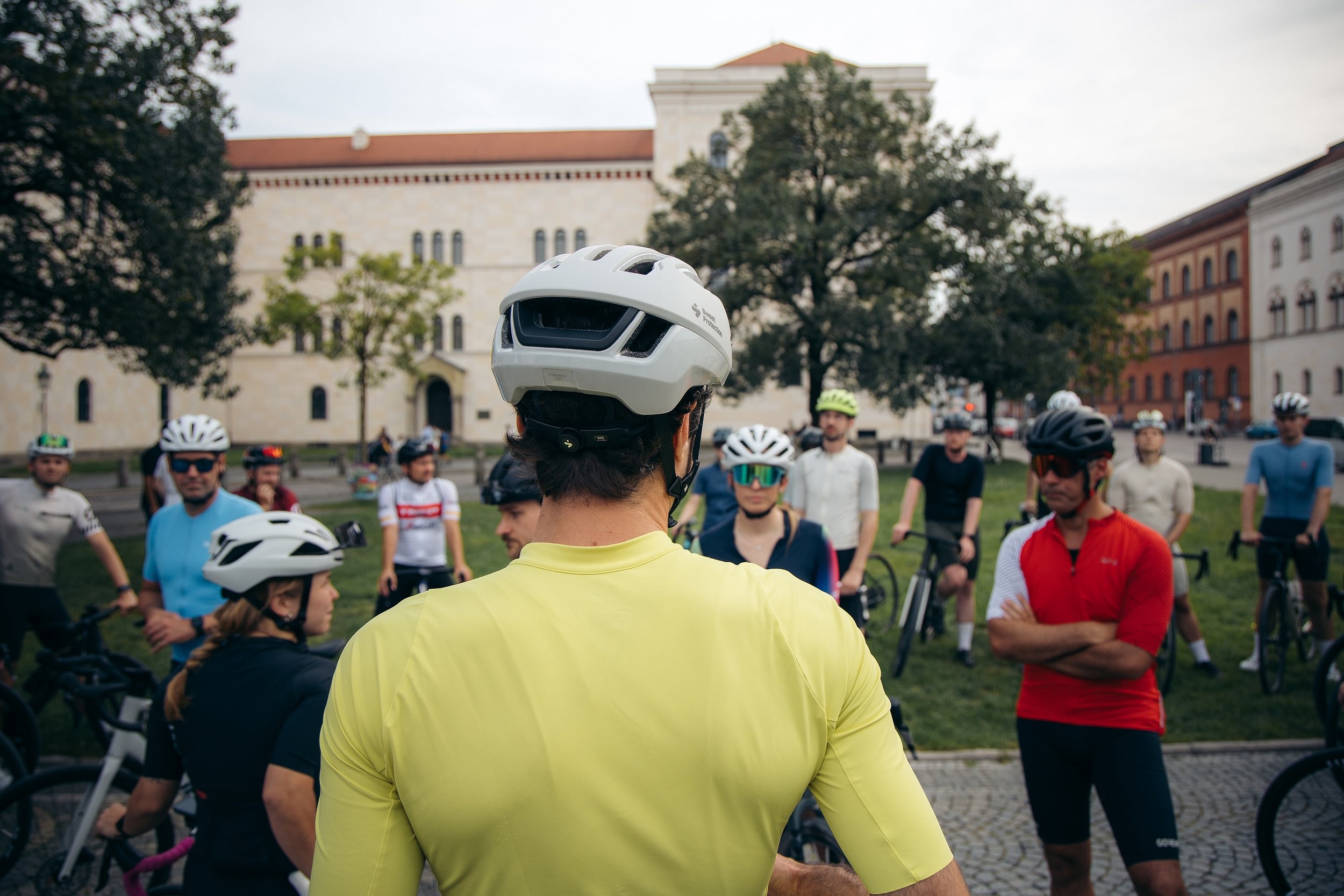 Eine Gruppe von Radfahrern, die sich vor einem historischen Gebäude versammelt haben, trägt Helme und Sportkleidung, eine Person in Gelb steht im Vordergrund.