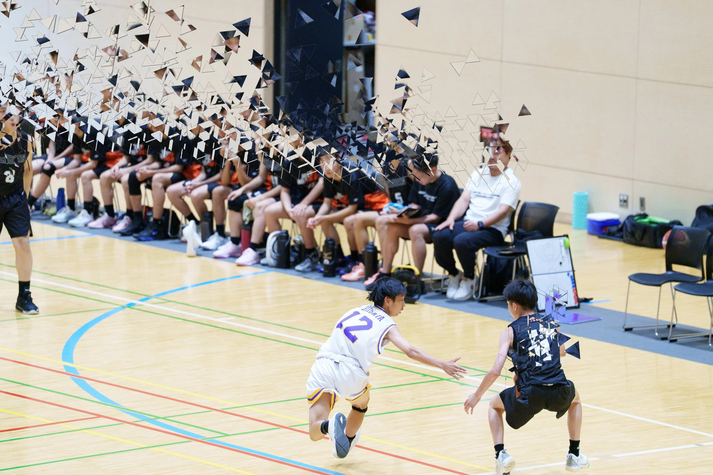 バスケットボールの試合中、コートでPlayer 同士が競り合っています。ベンチには複数の選手とコーチが座っていて、その瞬間を見つめ、応援しています。