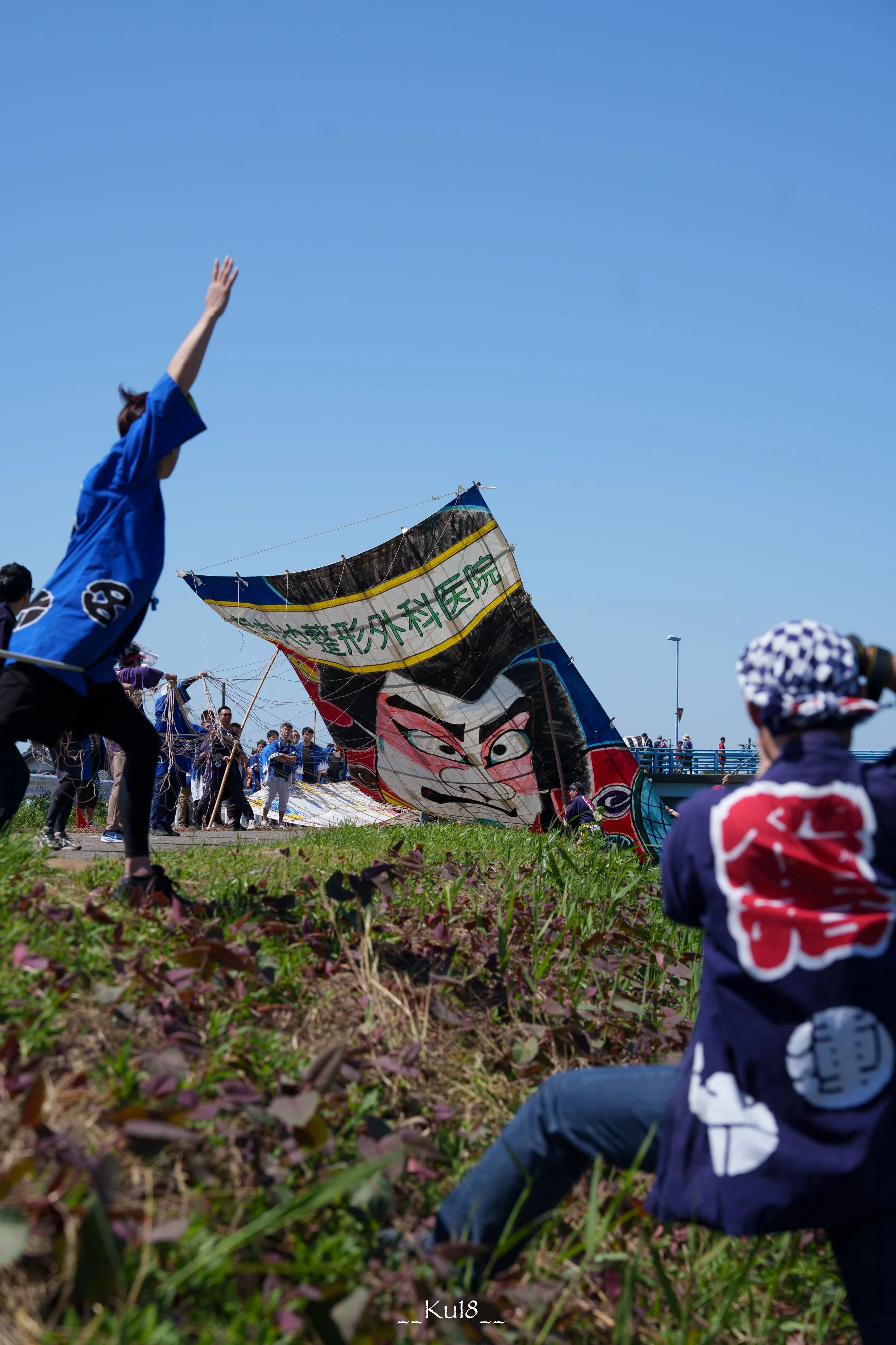 地面に座って、凧揚げをしている人々。青空の下で大きな凧が風に乗って空に上がろうとしている。