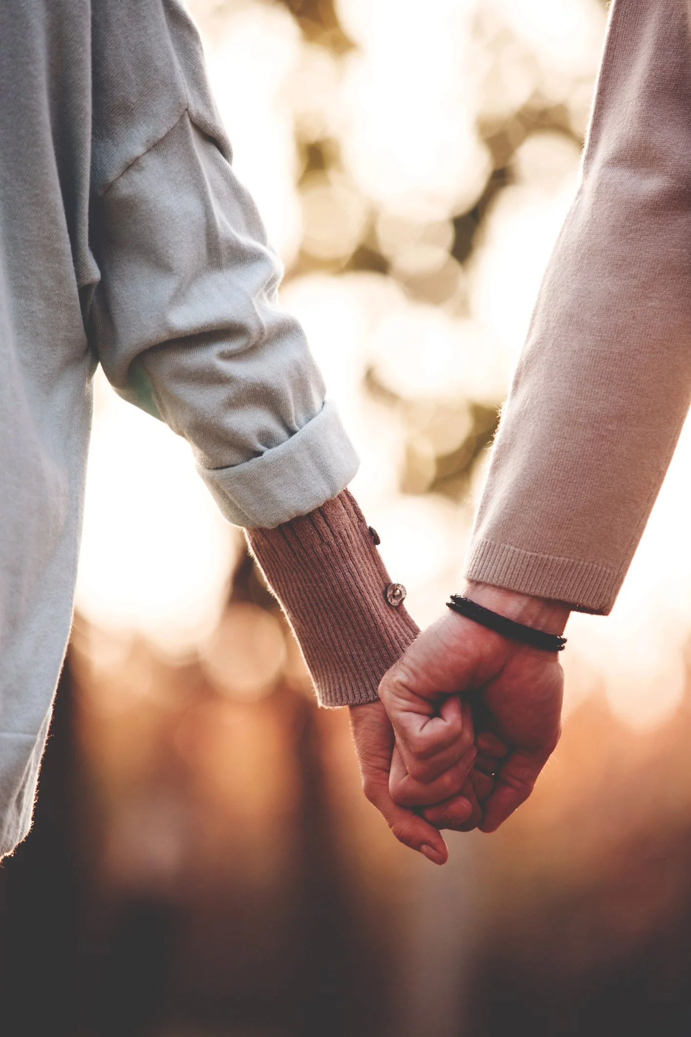 Close-up of two people holding hands outdoors, with blurred background of trees and sunlight.