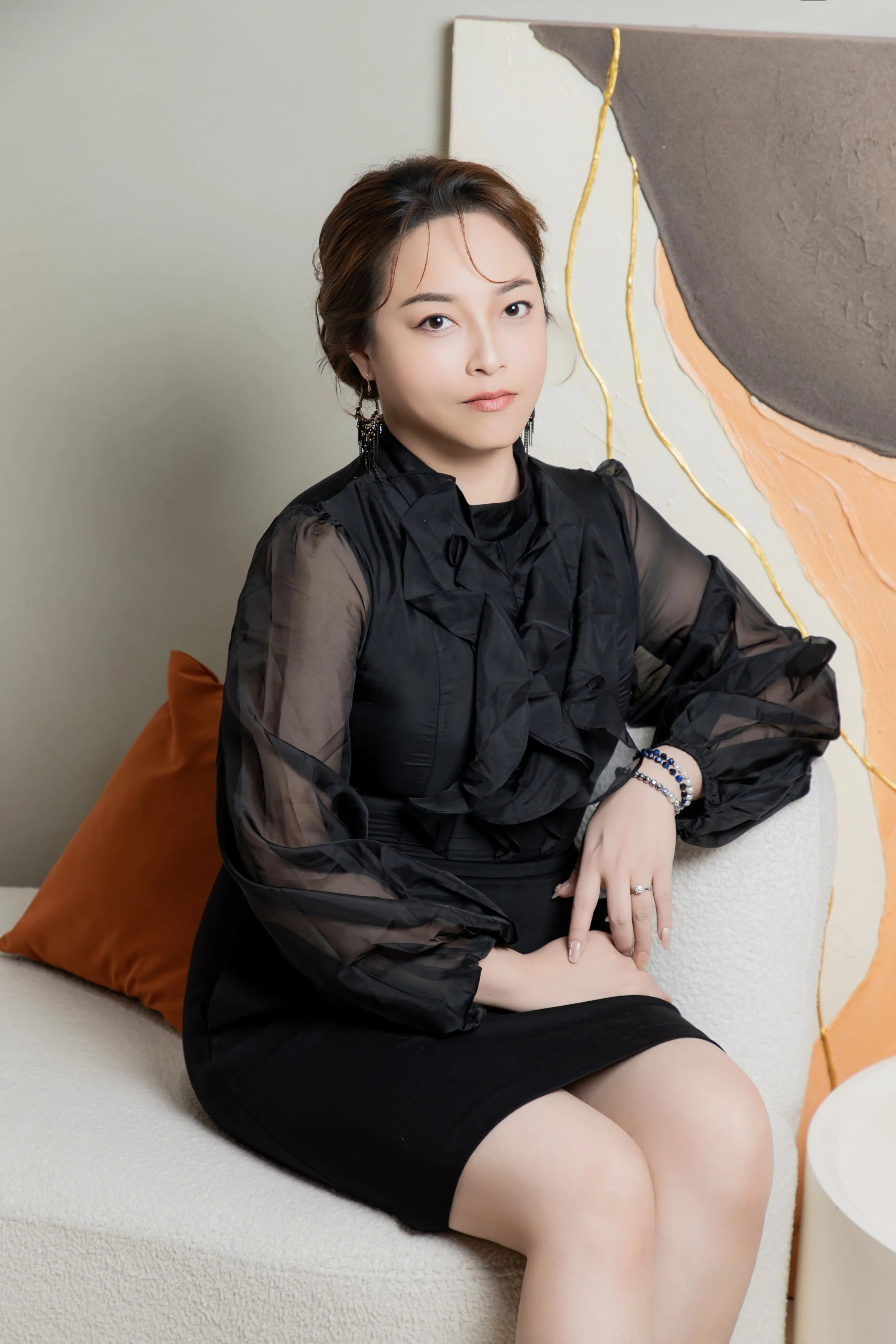 A woman with short brown hair, wearing a black dress with sheer puffed sleeves, is sitting on a white textured sofa with a brown cushion behind her. She is looking at the camera with a neutral expression, adorned with earrings and bracelets. Behind her is an abstract wall art with black, beige, peach, and gold accents.