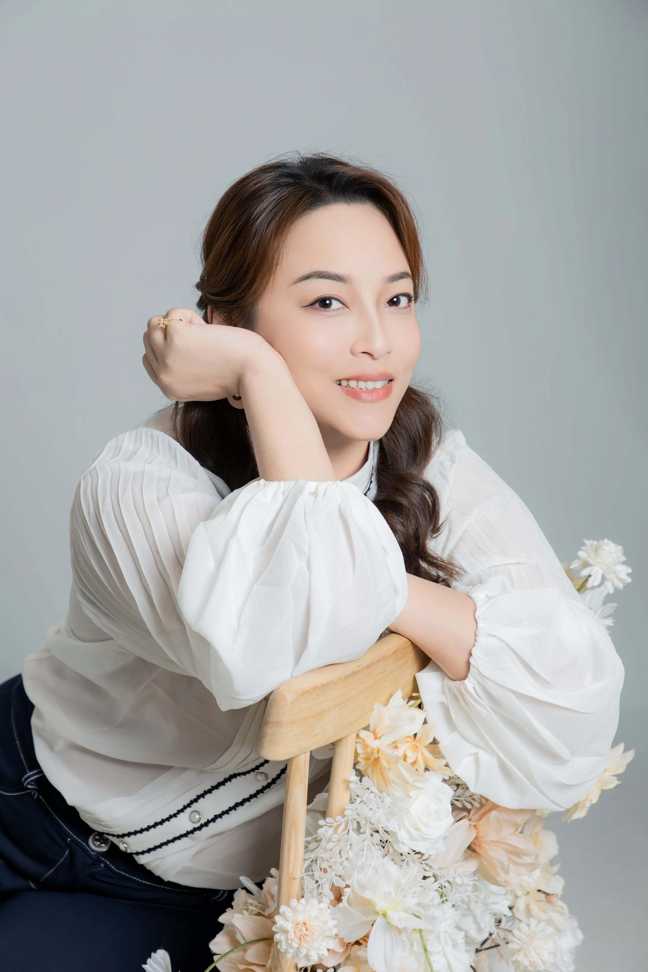 A woman with wavy brown hair smiling, resting her chin on her hand, leaning on a wooden chair decorated with white and peach flowers, against a plain background.