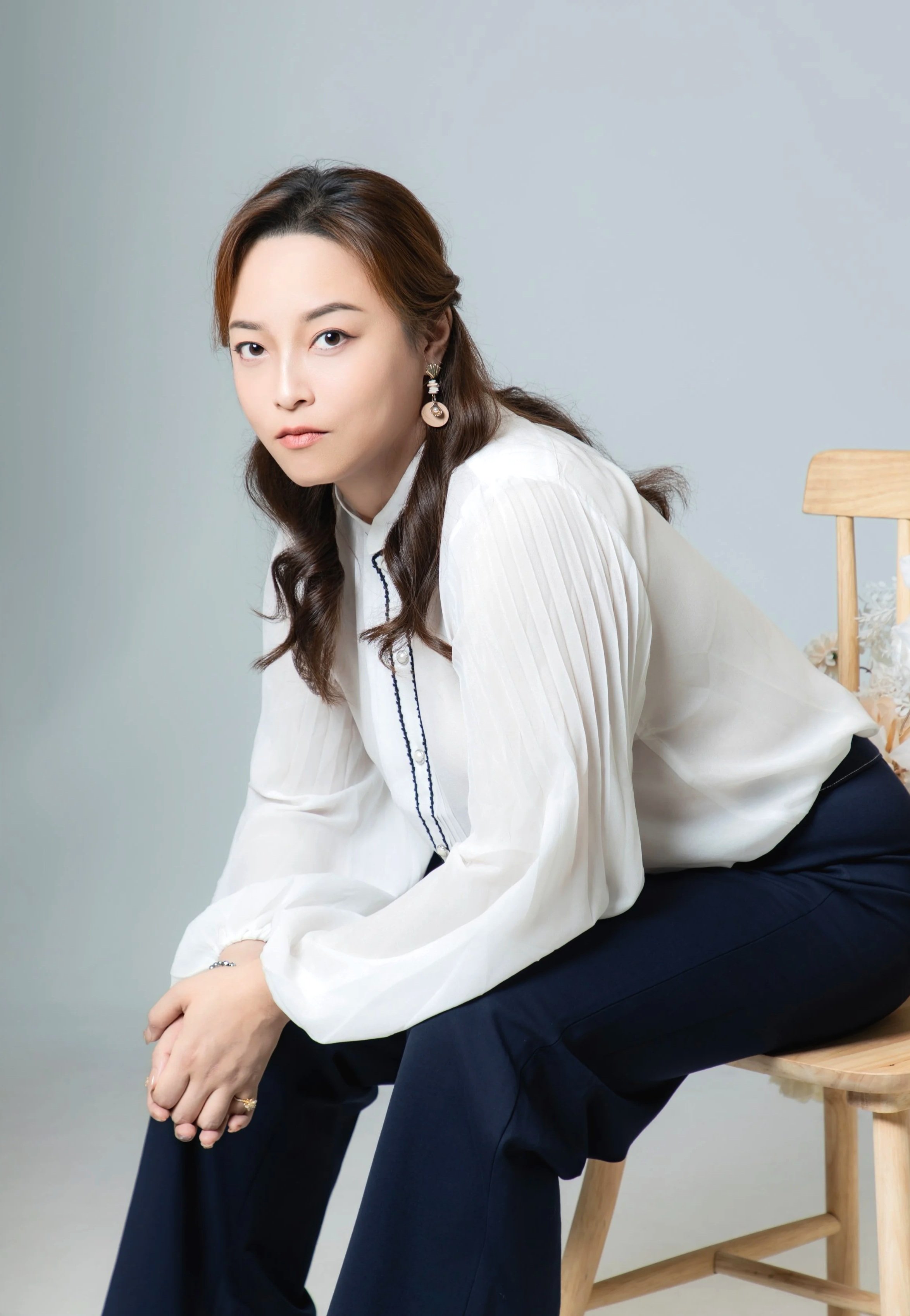 A woman with long, wavy brown hair sitting on a wooden chair, wearing a white blouse with pleated sleeves and dark pants, looking at the camera with a neutral expression.