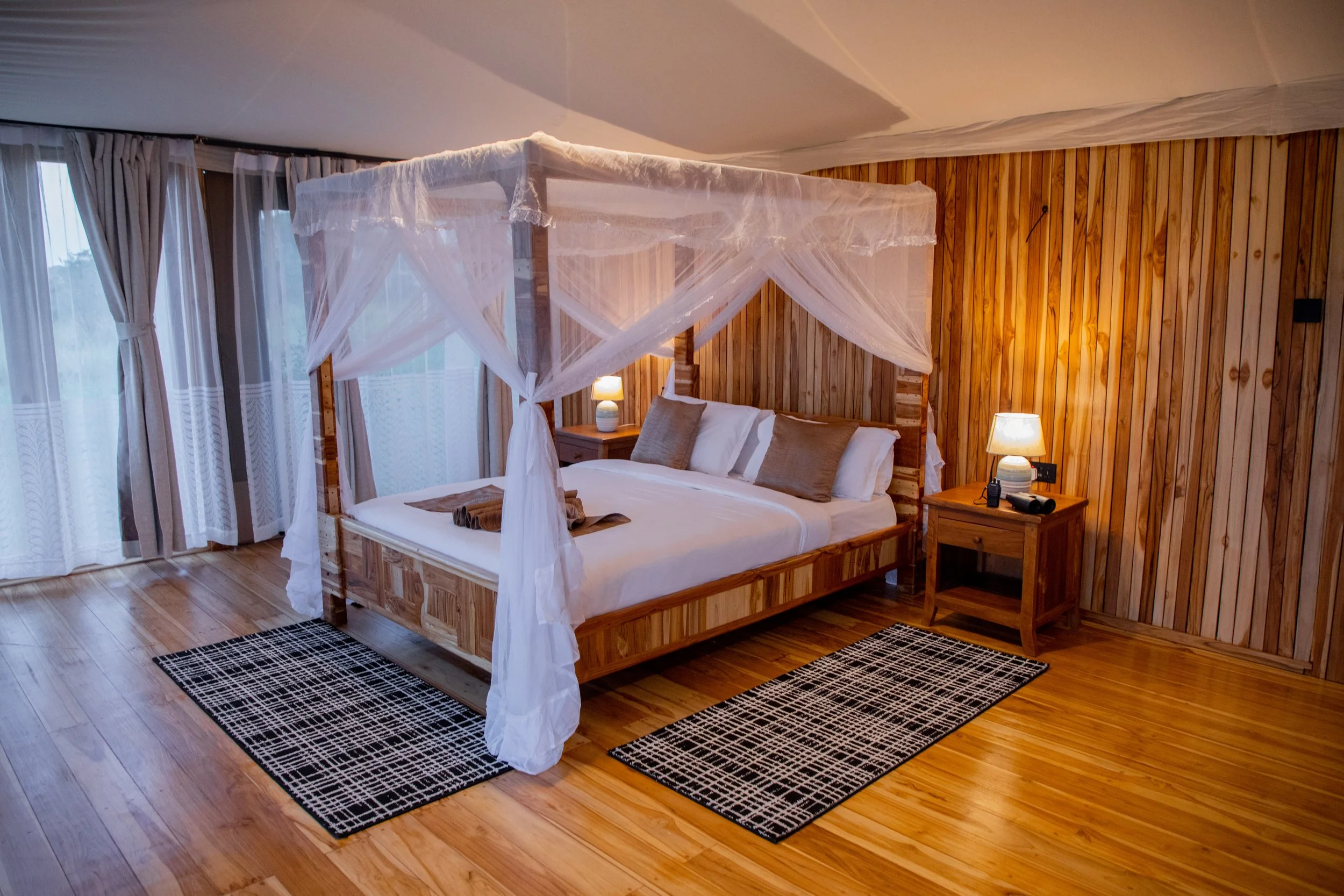 A cozy bedroom with a four-poster bed with white canopy, wooden headboard, and side tables. Two lamps on the side tables are illuminated. Large windows with sheer and heavy curtains are on the left, and the room has wooden floors and wall paneling wi