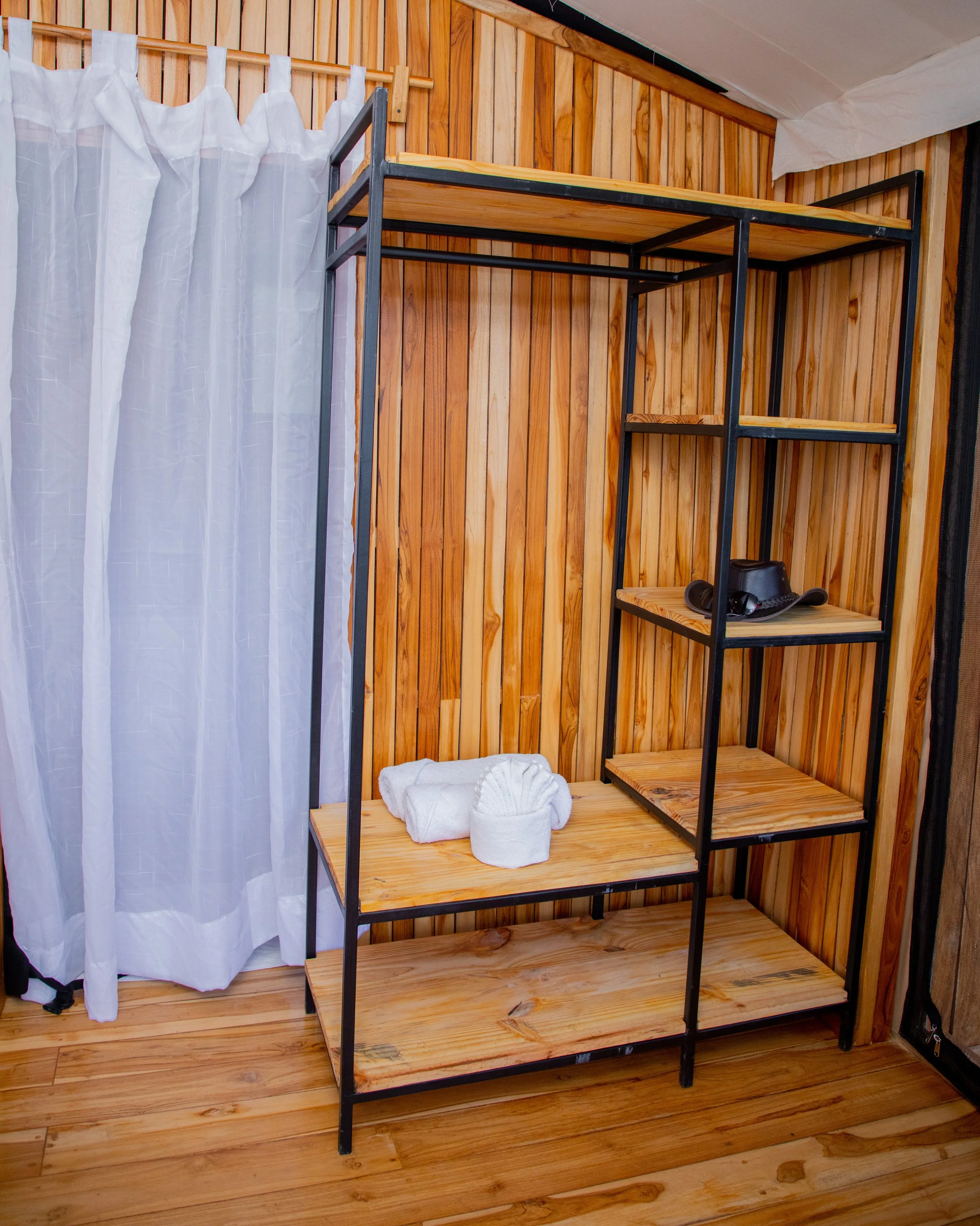 Wooden and black metal shelving unit with towels and toiletries, next to a white curtain and wood-paneled wall.