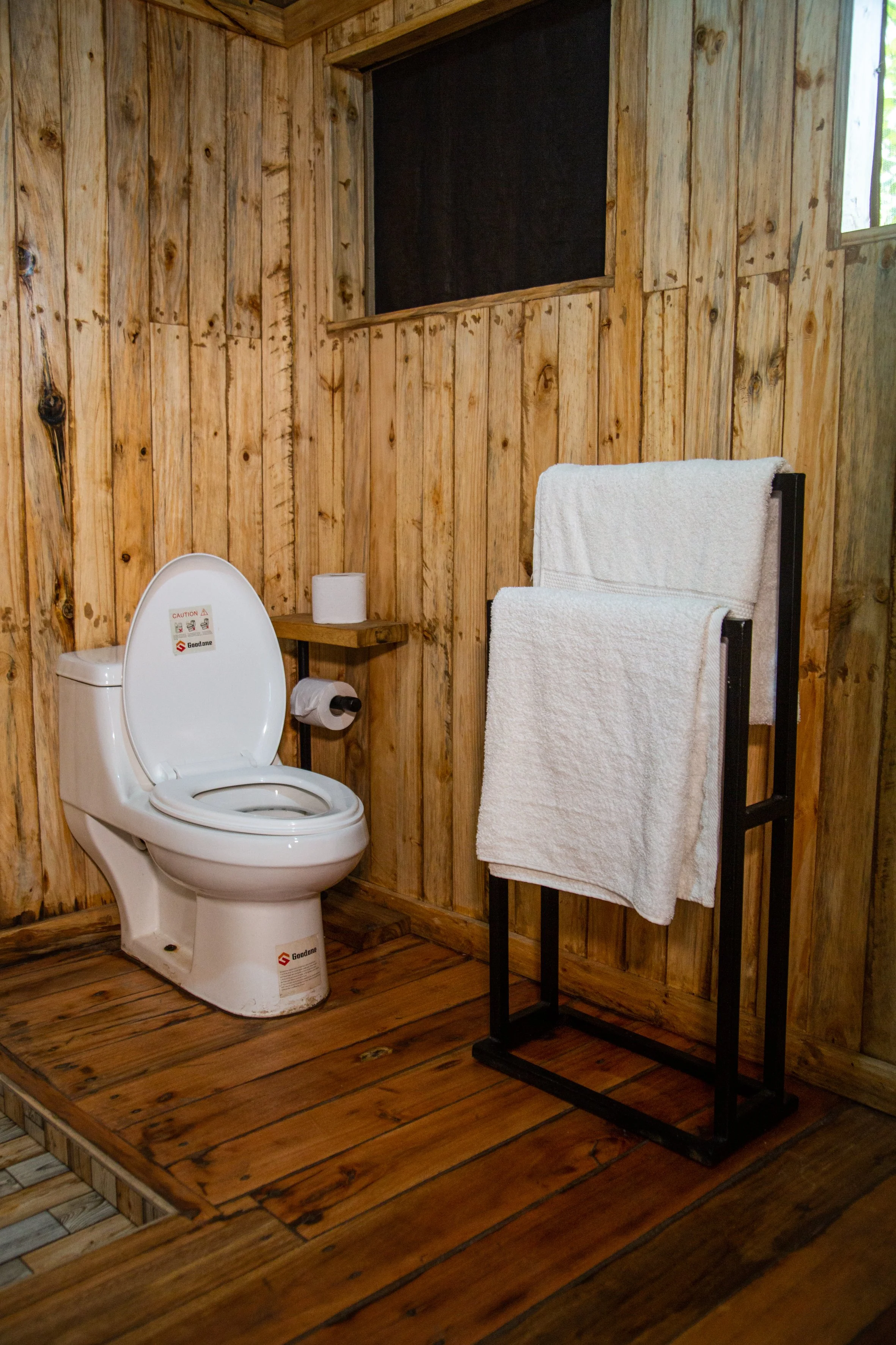 A rustic bathroom with wooden walls and floor, featuring a white toilet, a small wooden shelf holding toilet paper, and a black towel rack with white towels.