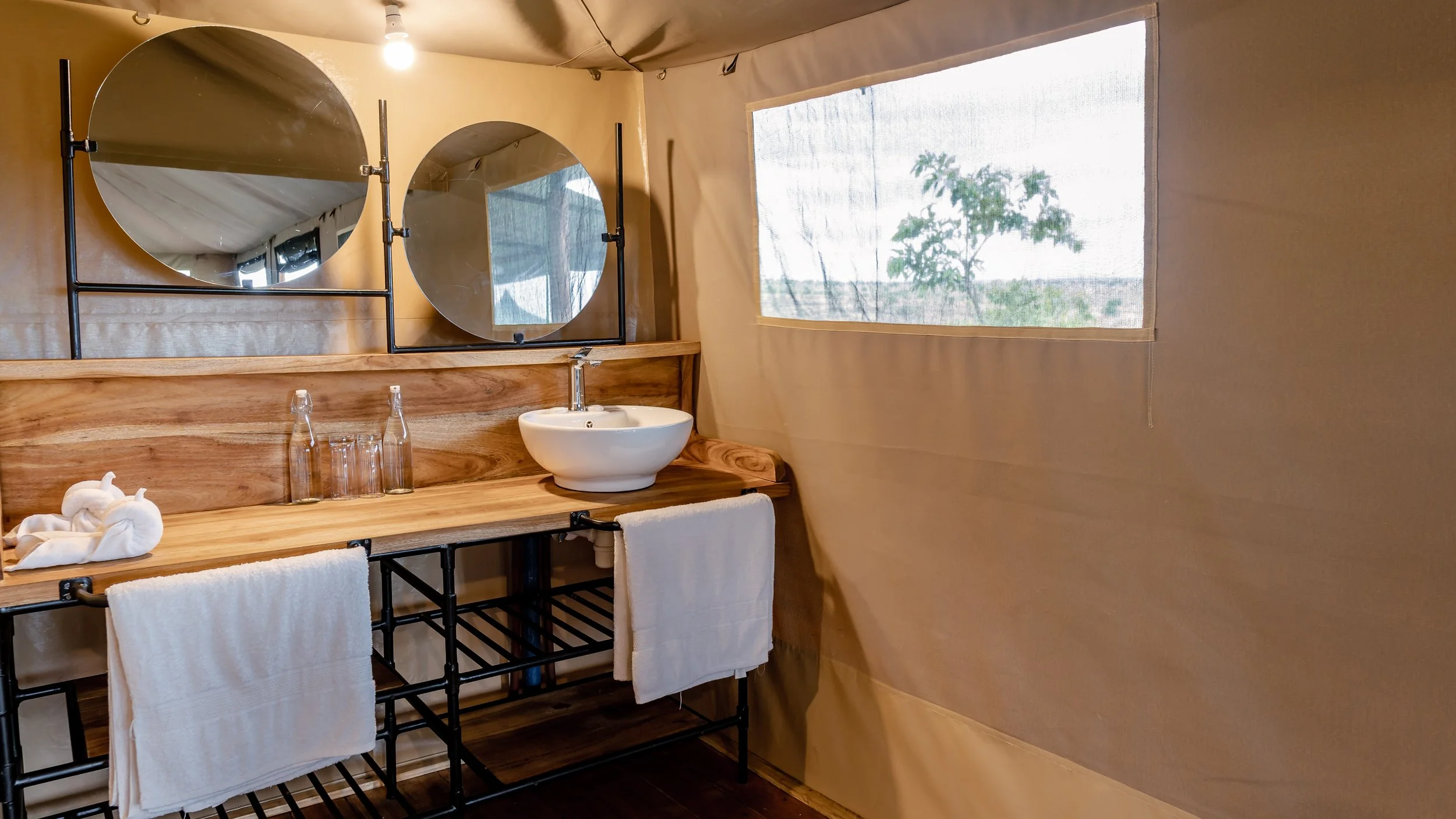 A rustic bathroom with a wooden vanity, two round mirrors, a small vessel sink, and two glass water bottles. Towels hang on a black metal towel rack beneath the vanity, and a large window with a view of trees is on the right side.