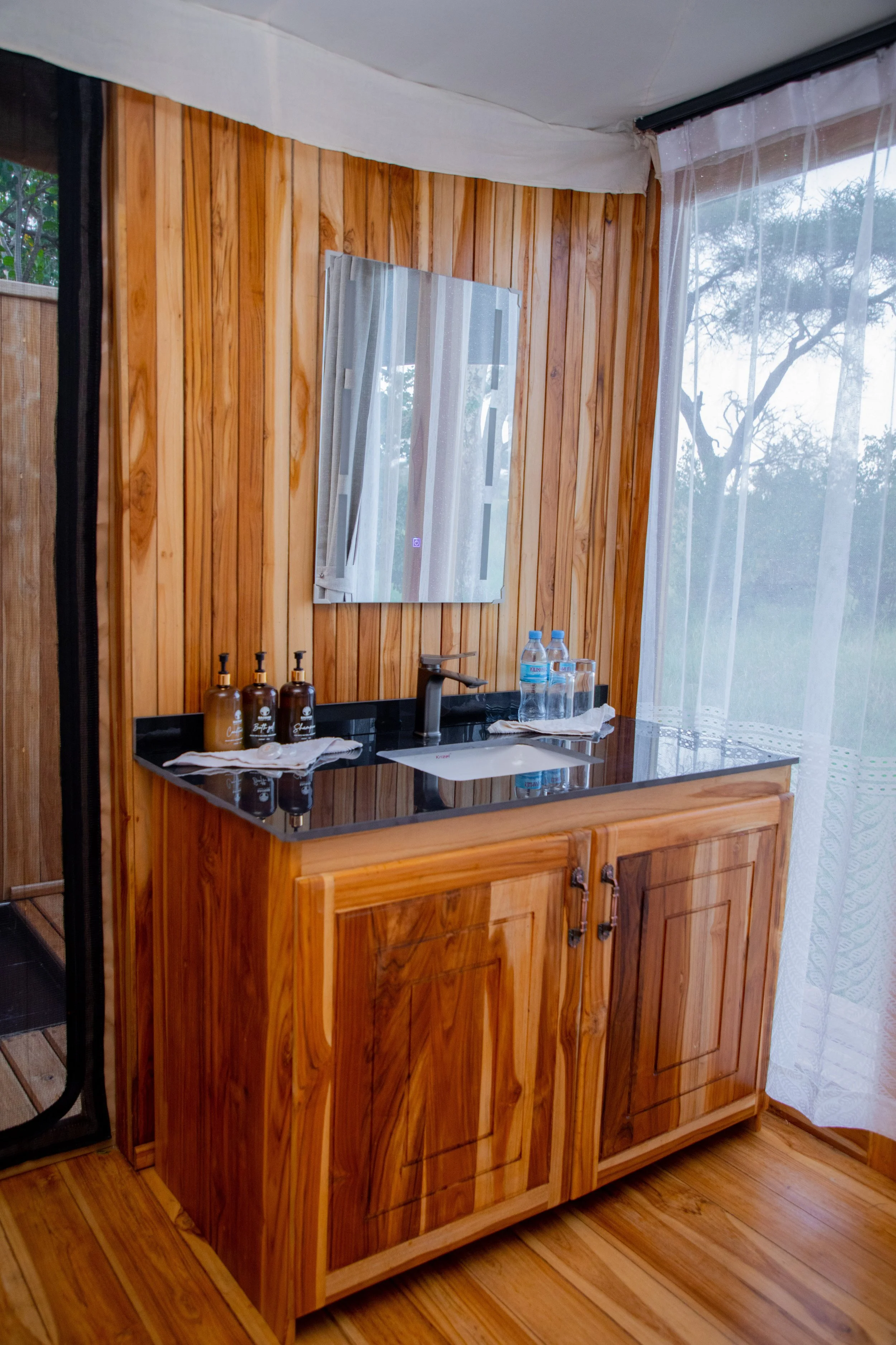 Wooden bathroom vanity with a black countertop, a small sink, and a mirror. Bottles of soap and water bottles are on the counter. There are white curtains on a large window next to the vanity.