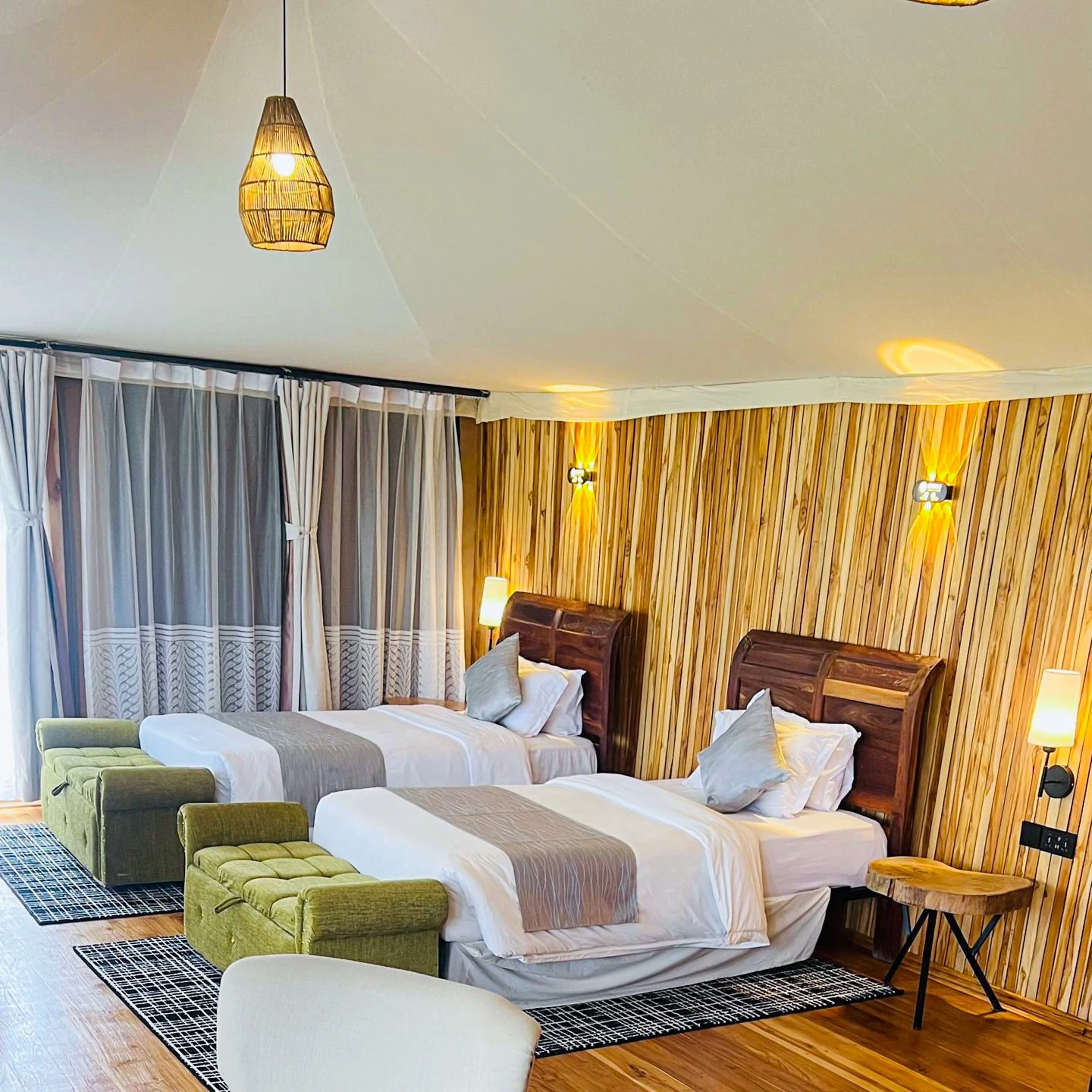 A hotel room with two single beds featuring wooden headboards, gray and white bedding, and decorative pillows. Each bed has a matching ottoman at the foot, one green and one beige. The room has a wooden accent wall, wall-mounted lights, and a floor l