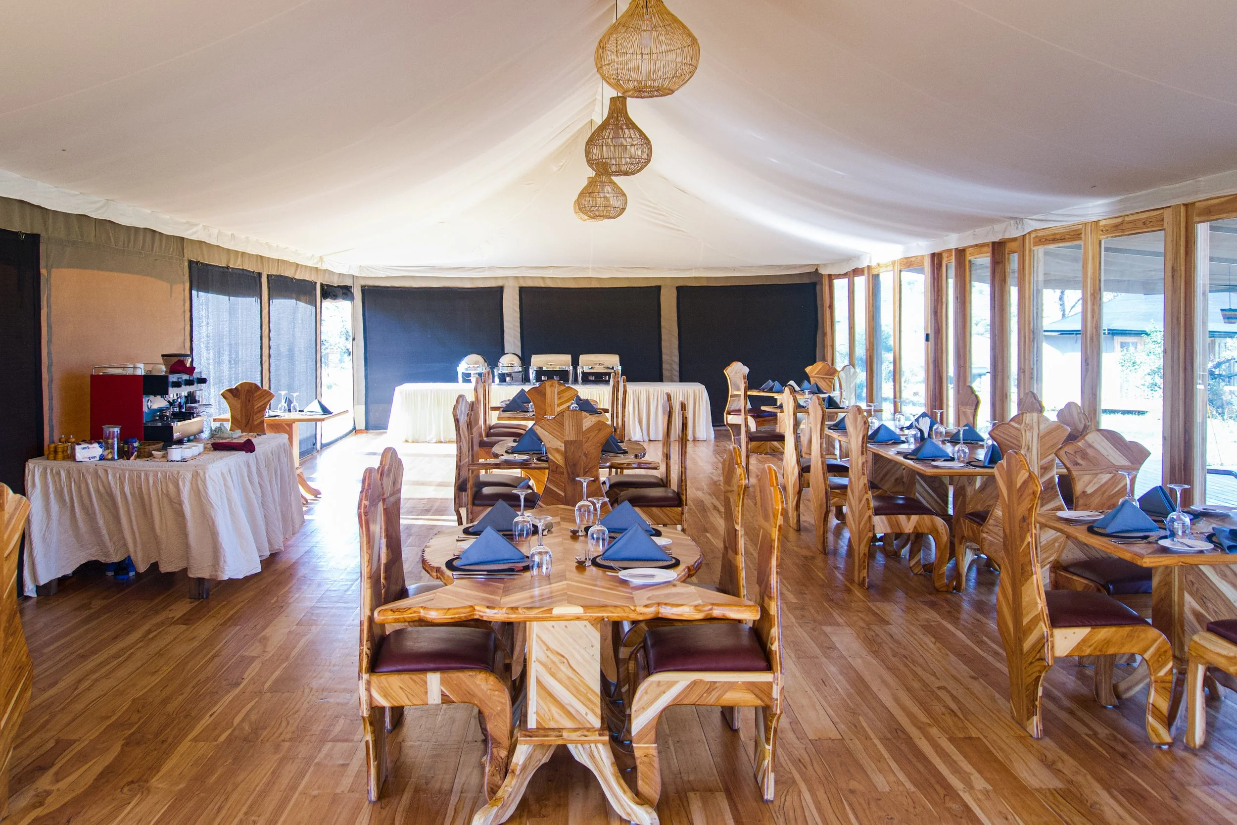 Elegant dining room set up with wooden tables and chairs, blue napkins, glassware, and a buffet area with coffee and tea station, in a bright room with large windows and hanging pendant lights.