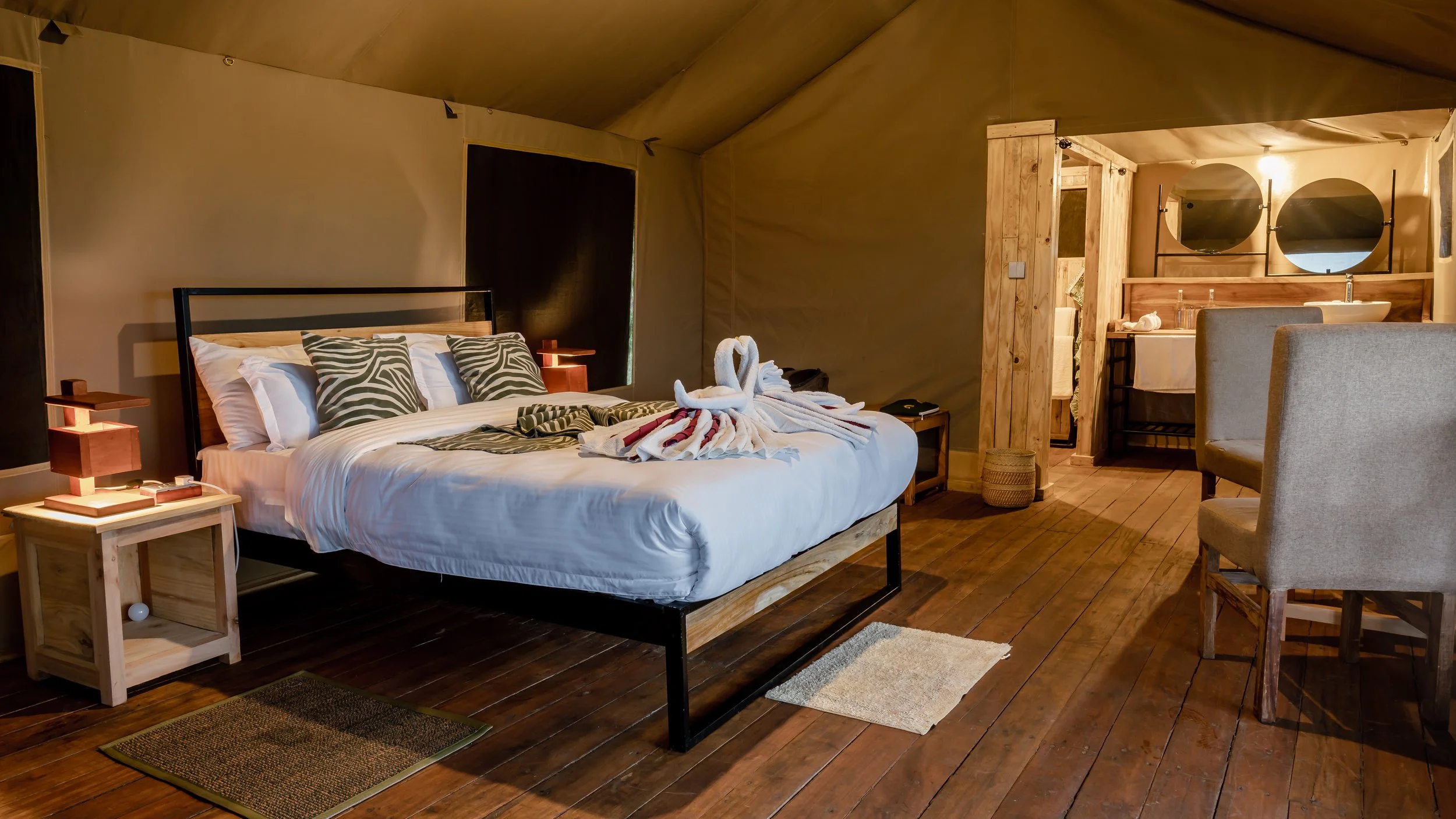 A cozy bedroom with a large bed decorated with white linens and two patterned pillows, a towel art in the shape of a swan, wooden nightstands, a seating area with chairs, and a bathroom area visible in the background with a sink and vanity.
