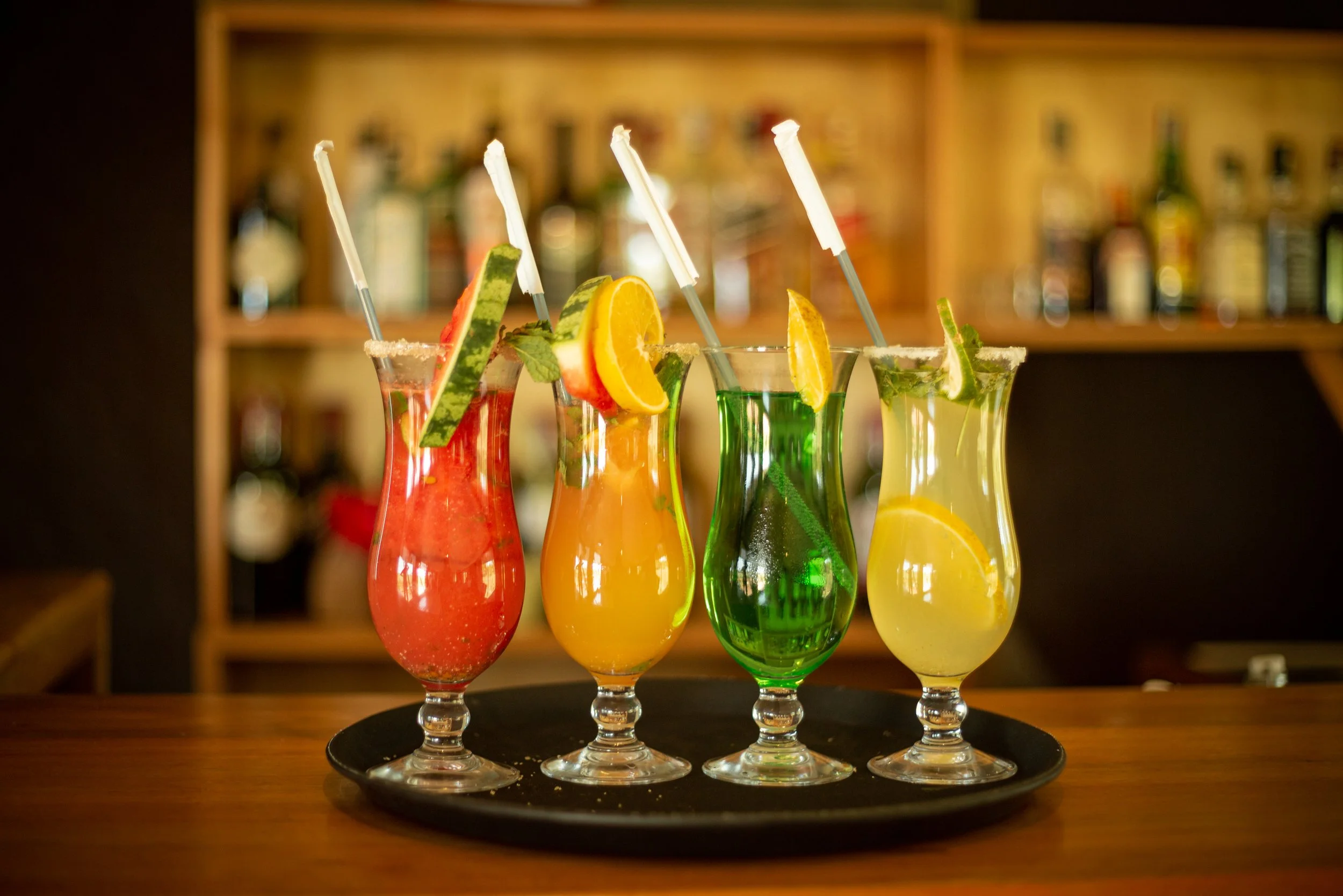 Four colorful cocktails with fruit garnishes and straws on a black tray on a wooden bar counter.