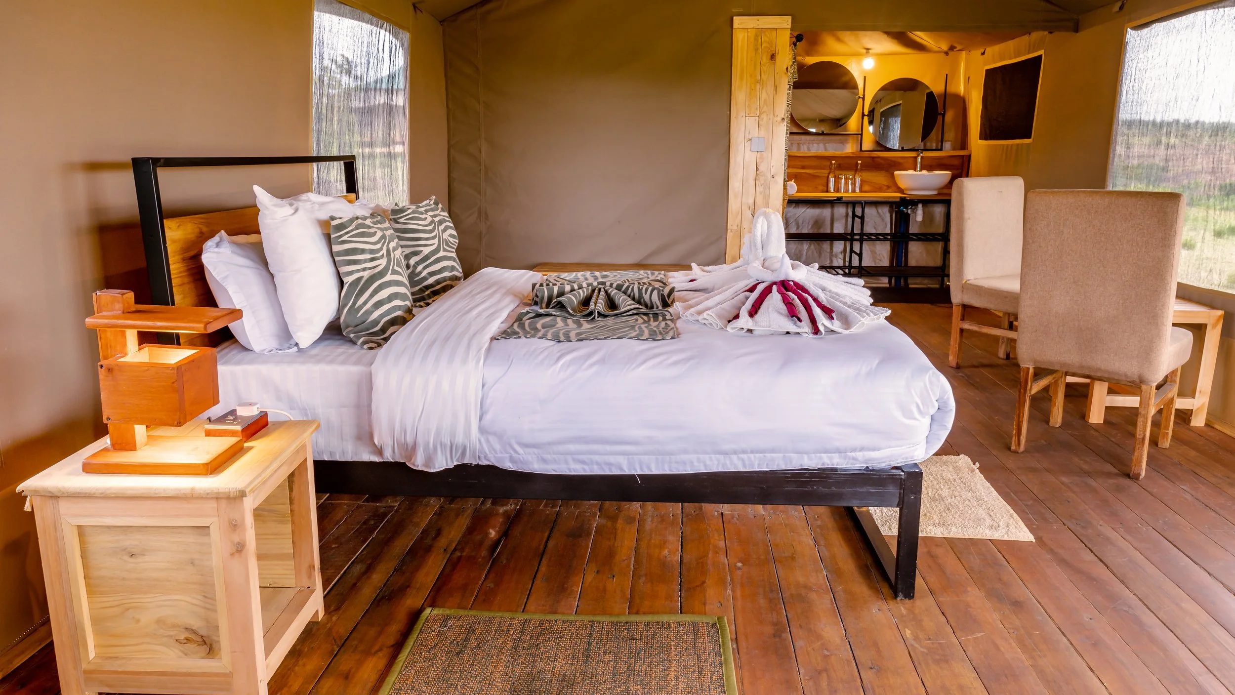 A cozy bedroom inside a tent with a large bed made with white sheets, patterned pillows, and towel art shaped like swans. There is a wooden nightstand with a lamp and a charger. In the background, the tent has a small bathroom sink and a mirror, and