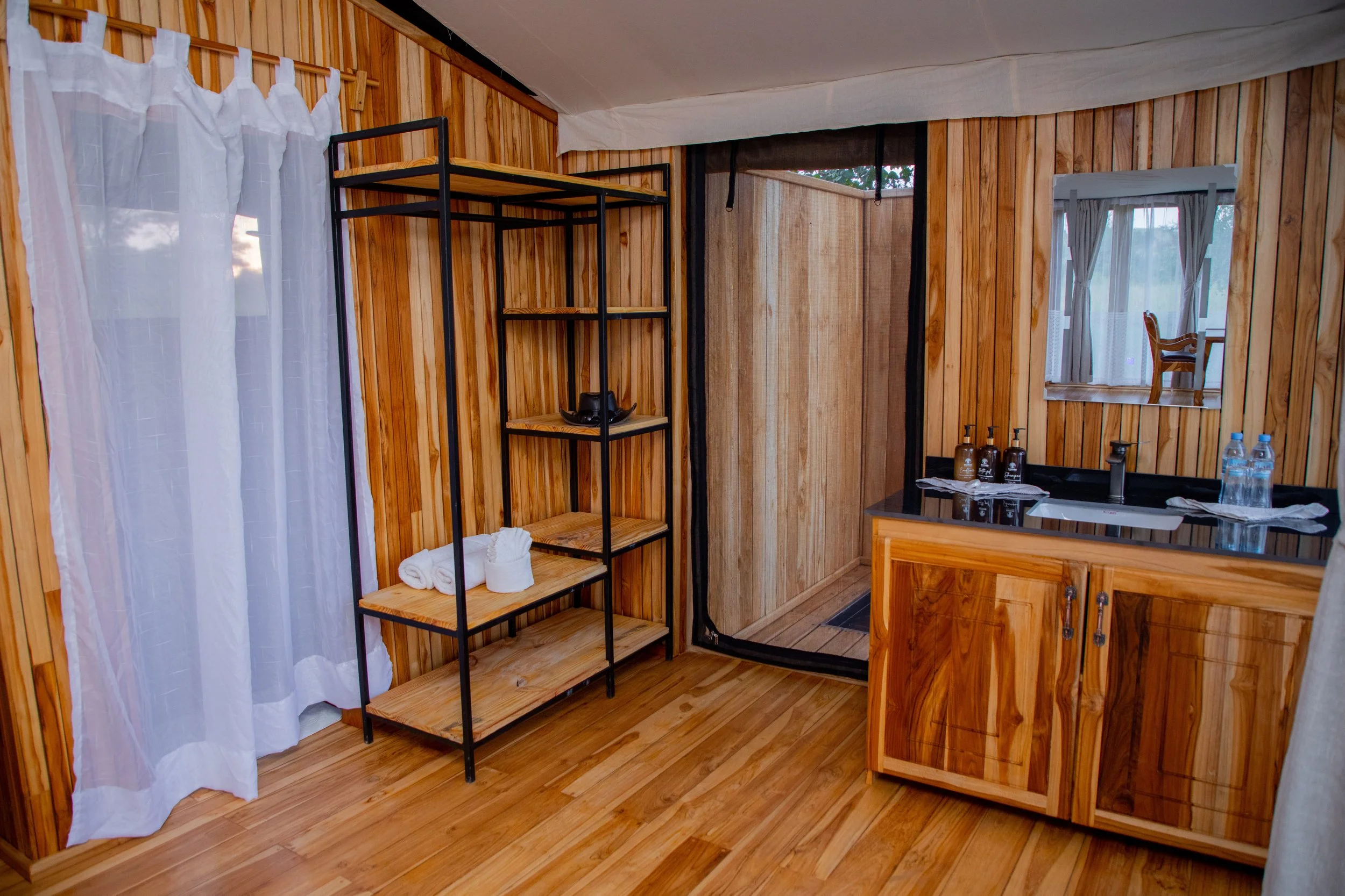 Interior of a rustic wooden bathroom with a shower, vanity with a black countertop, towels, toiletries, a mirror, and a small window.