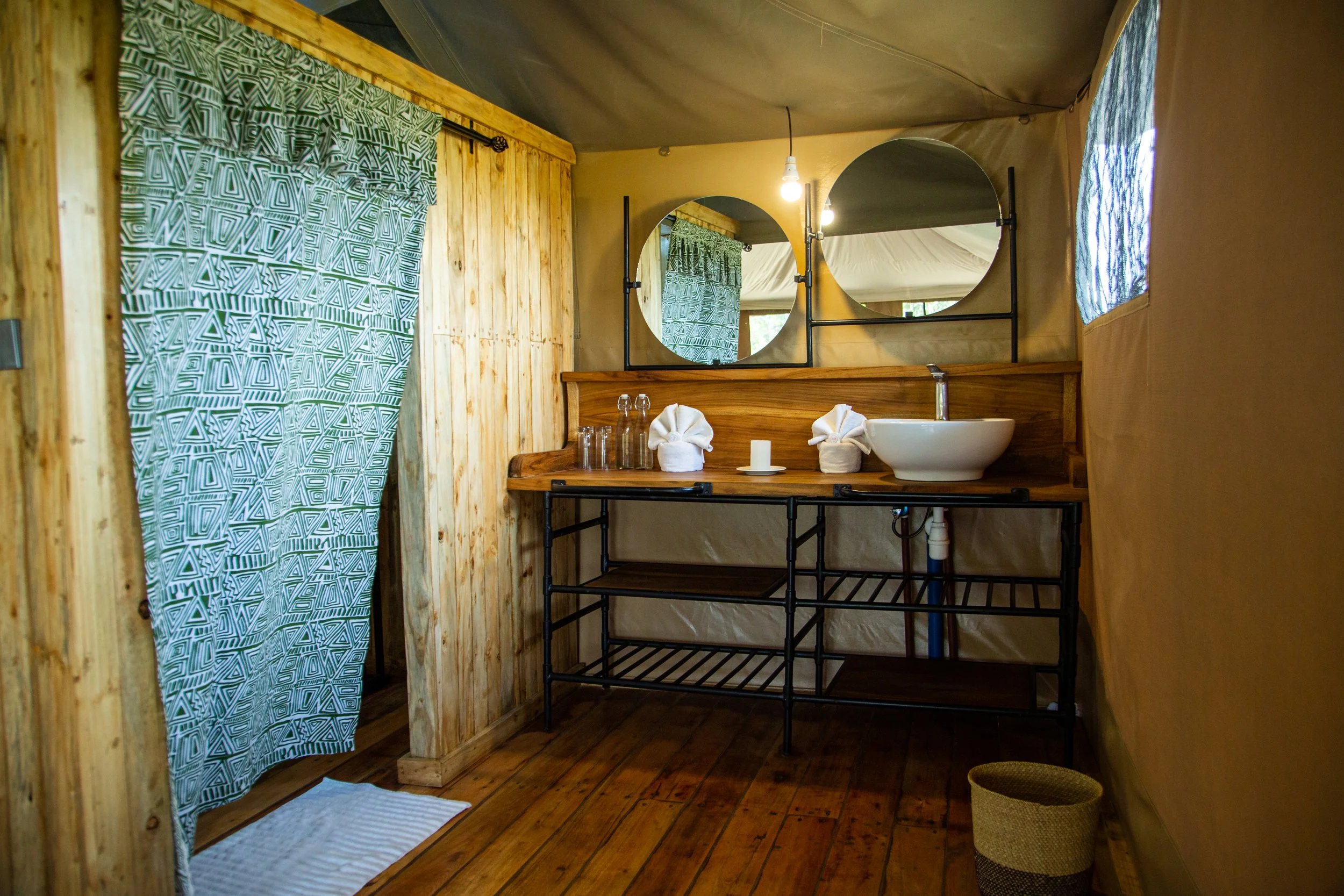 Rustic bathroom with wooden walls and floors, featuring a white vessel sink on a wooden and metal vanity, two round mirrors, and a window with patterned curtain.