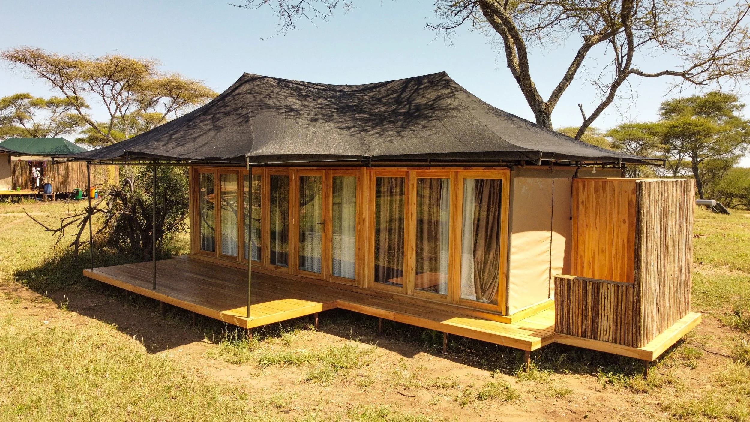 A wooden safari tent with glass doors, elevated on a wooden platform, surrounded by trees and grassland.