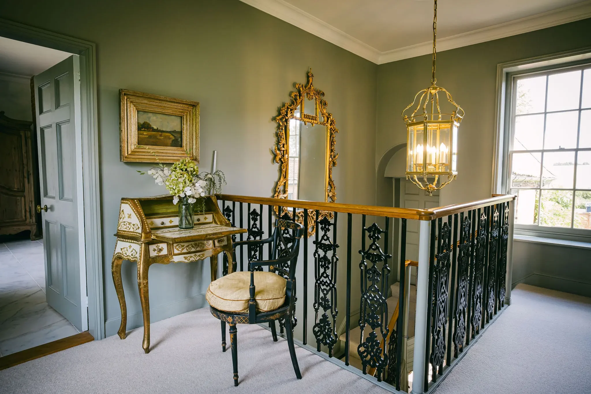 Interior of a house with a staircase, gold-framed mirror, landscape painting, a small gold and green table with a vase of flowers, and a decorative chandelier, with large windows allowing natural light.