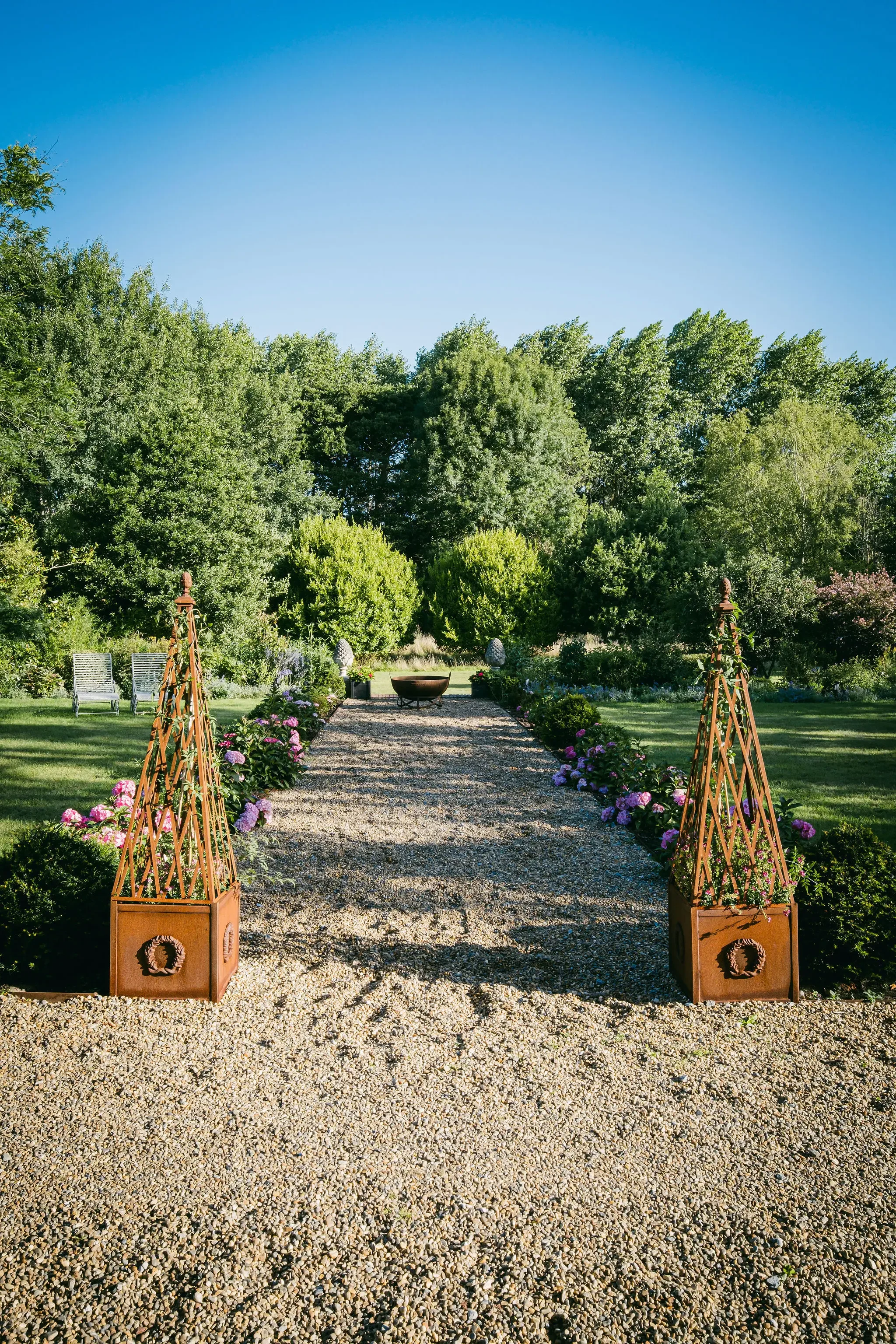 A gravel pathway in a landscaped garden with blooming pink and purple flowers on the sides, leading to a fire pit and surrounded by trees under a clear blue sky.