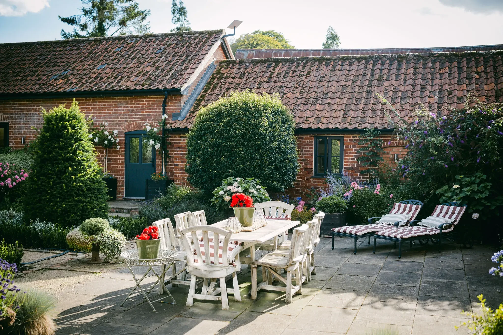 A brick house with a tile roof and a backyard garden featuring outdoor furniture, flowers, and lush greenery.