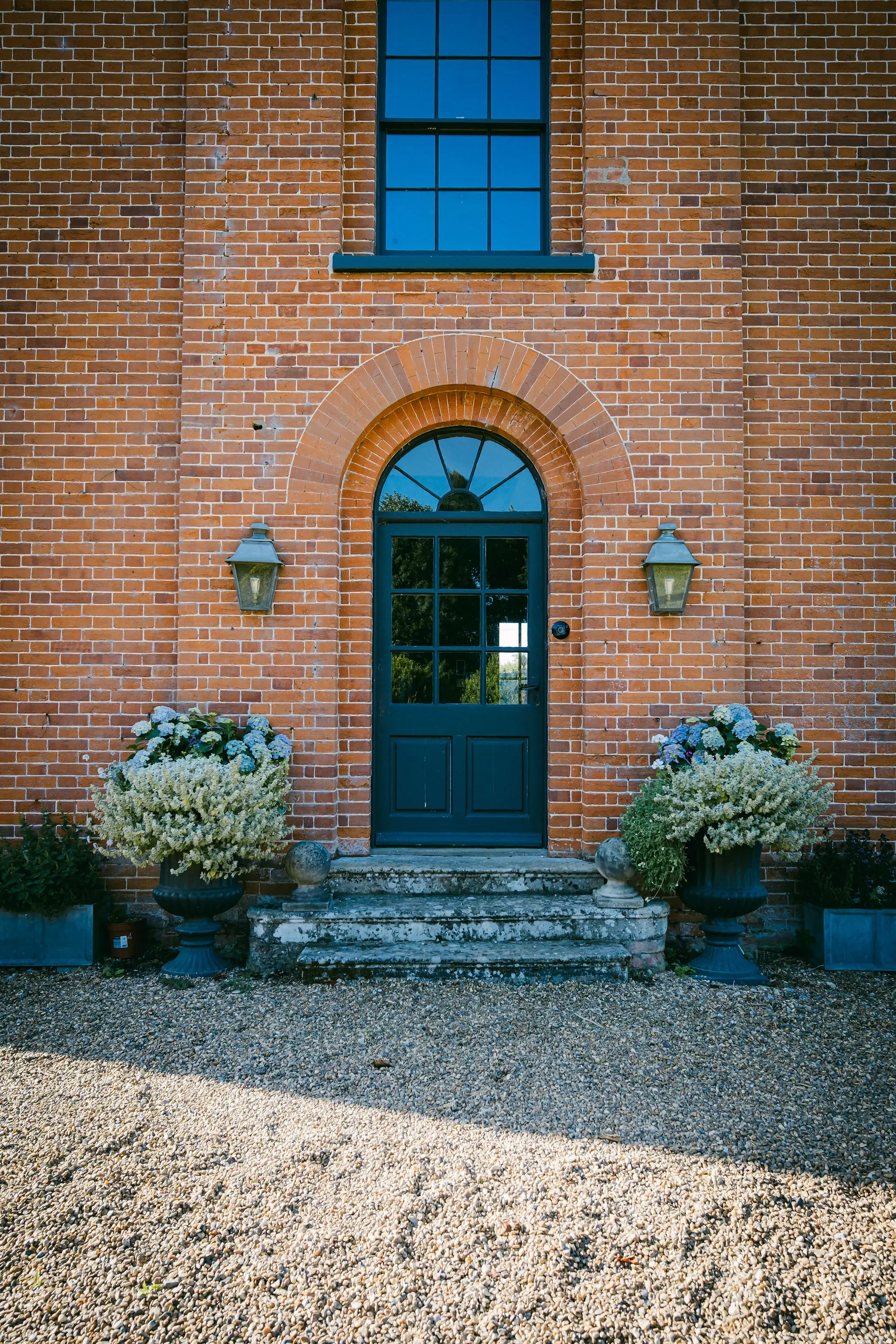 A brick house facade with a navy blue front door and window, flanked by two large planters with white flowers and greenery, and two outdoor wall lanterns.