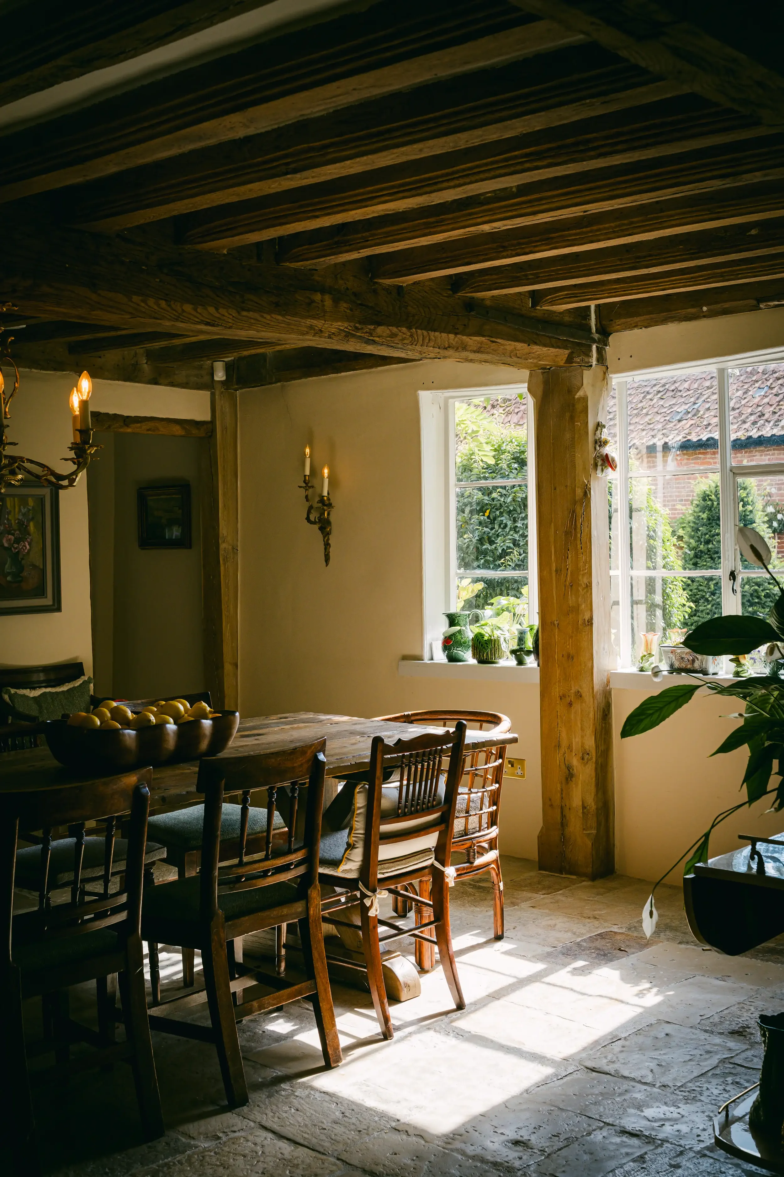 A rustic kitchen or dining room with wooden beams on the ceiling, a wooden dining table with chairs, natural light coming through large windows, and potted plants on the windowsills.