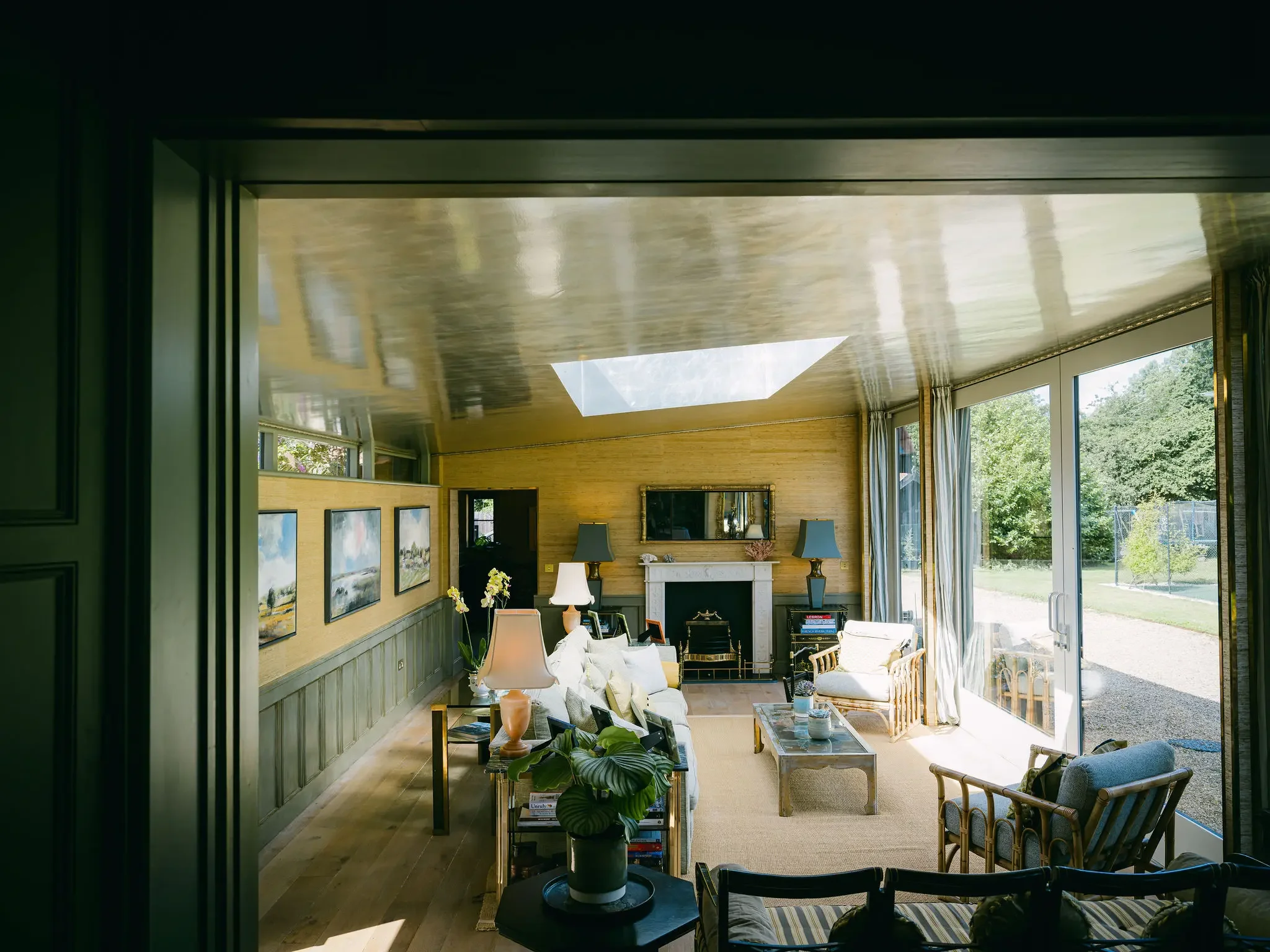 A spacious living room with large floor-to-ceiling windows, a fireplace, wall art, and various cushions and furniture, with a view of greenery outside.