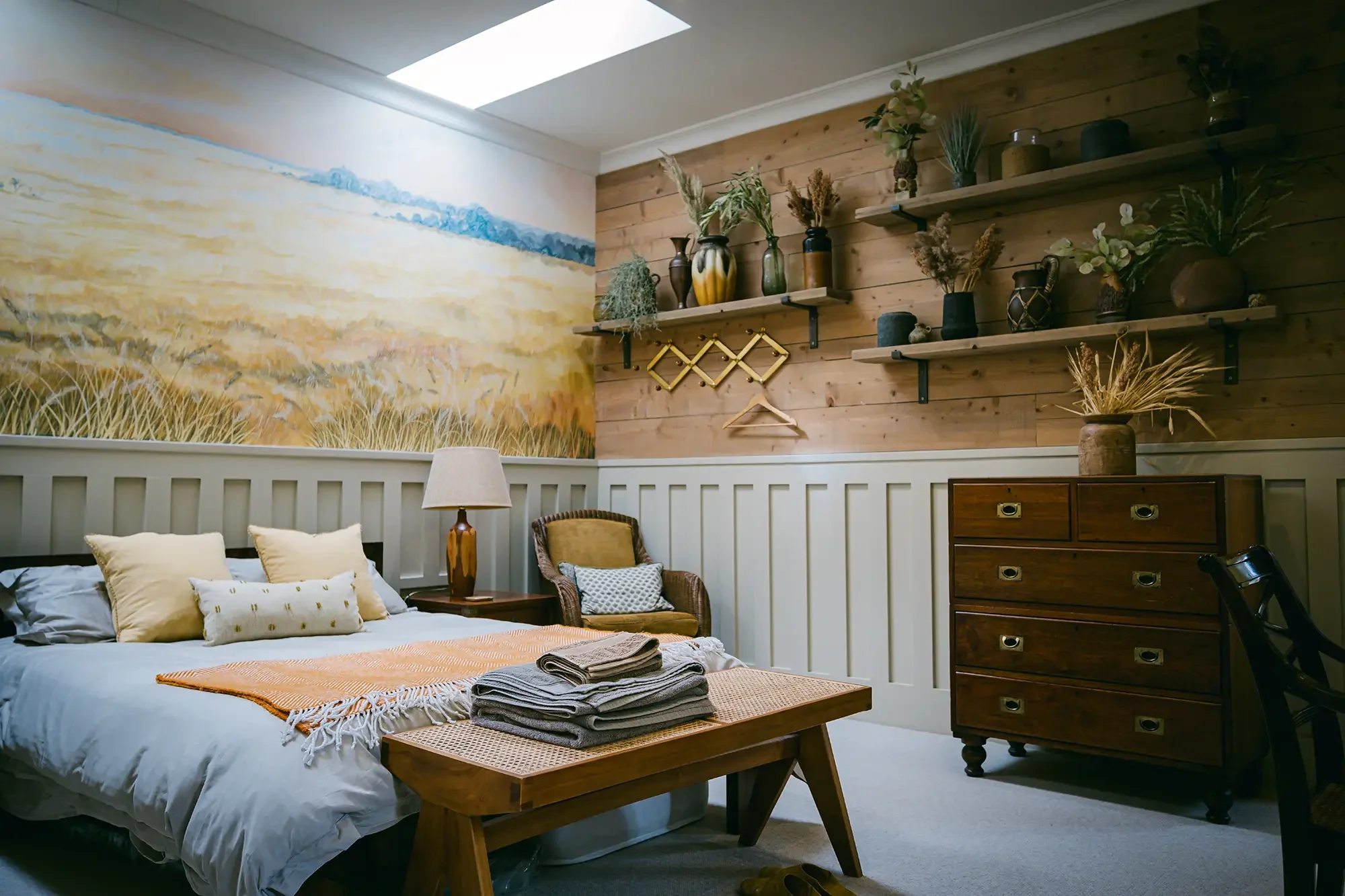 A cozy bedroom featuring a bed with beige and white pillows, a bedside lamp, and a wooden nightstand. There are folded towels on a wooden bench at the foot of the bed. The room has a decorative mural of a field and sky on one wall, and a wooden accent wall with shelves holding various vases and plants.