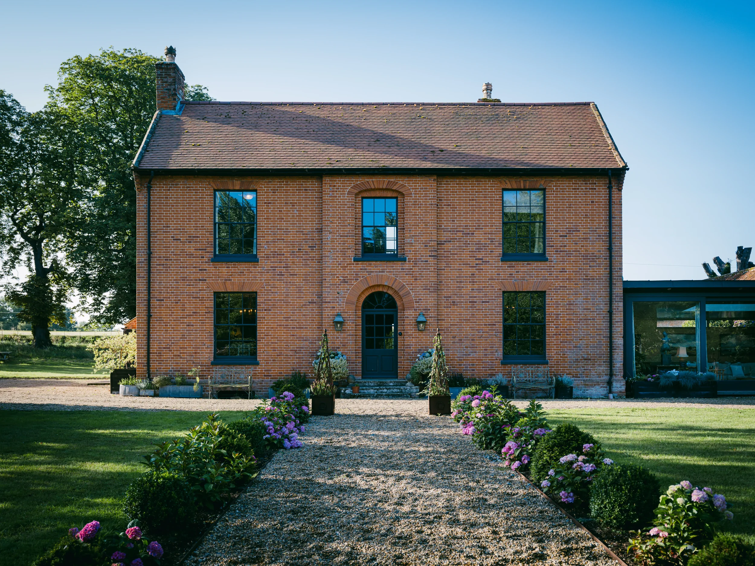 Front view of a two-story brick house with black window frames and a black front door, surrounded by a landscaped yard with pink and purple flowers and a gravel pathway.