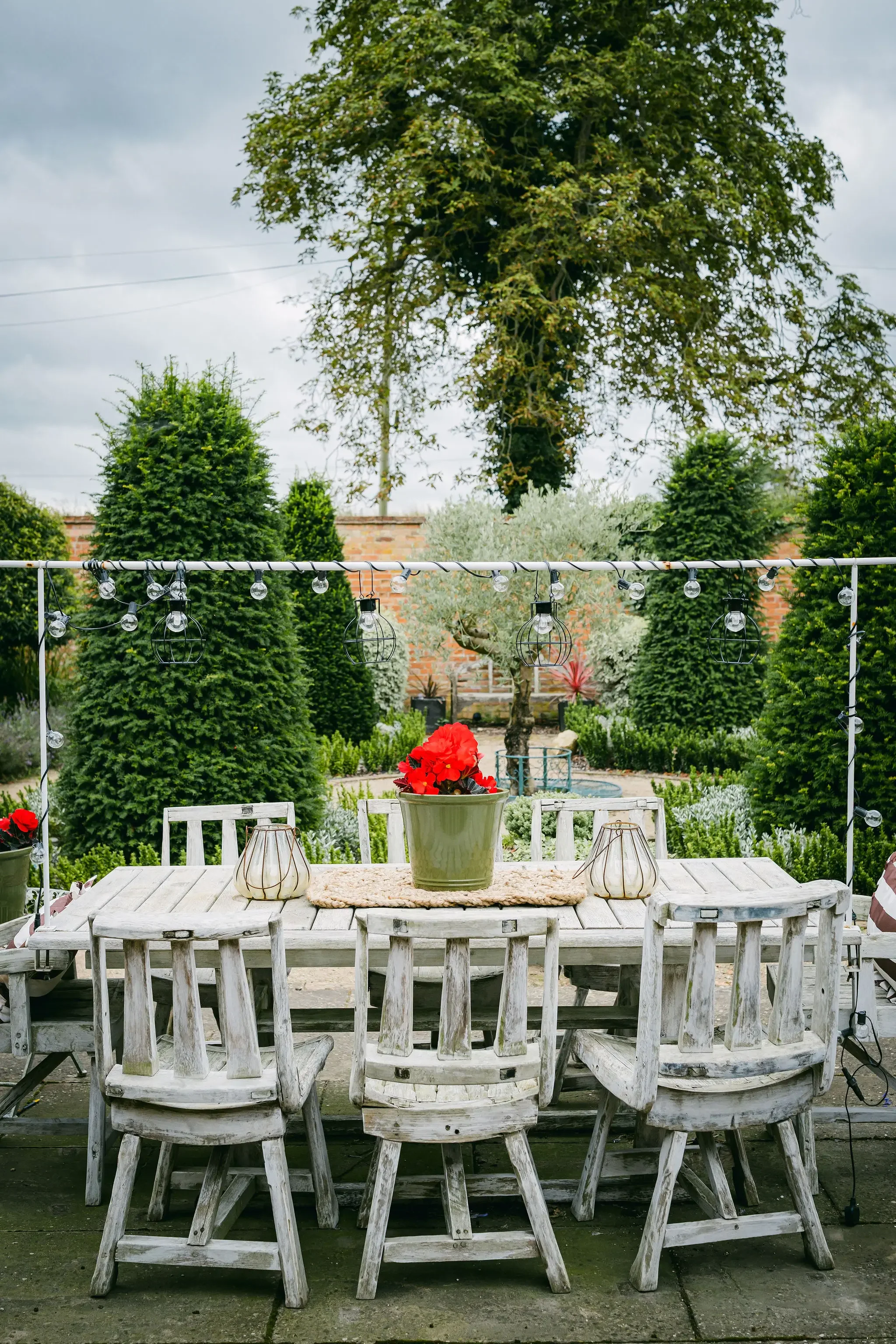 A white outdoor table with matching chairs, a green potted plant with red flowers, and two lanterns on a beige mat. In the background, there are tall green bushes, trees, and a cloudy sky.