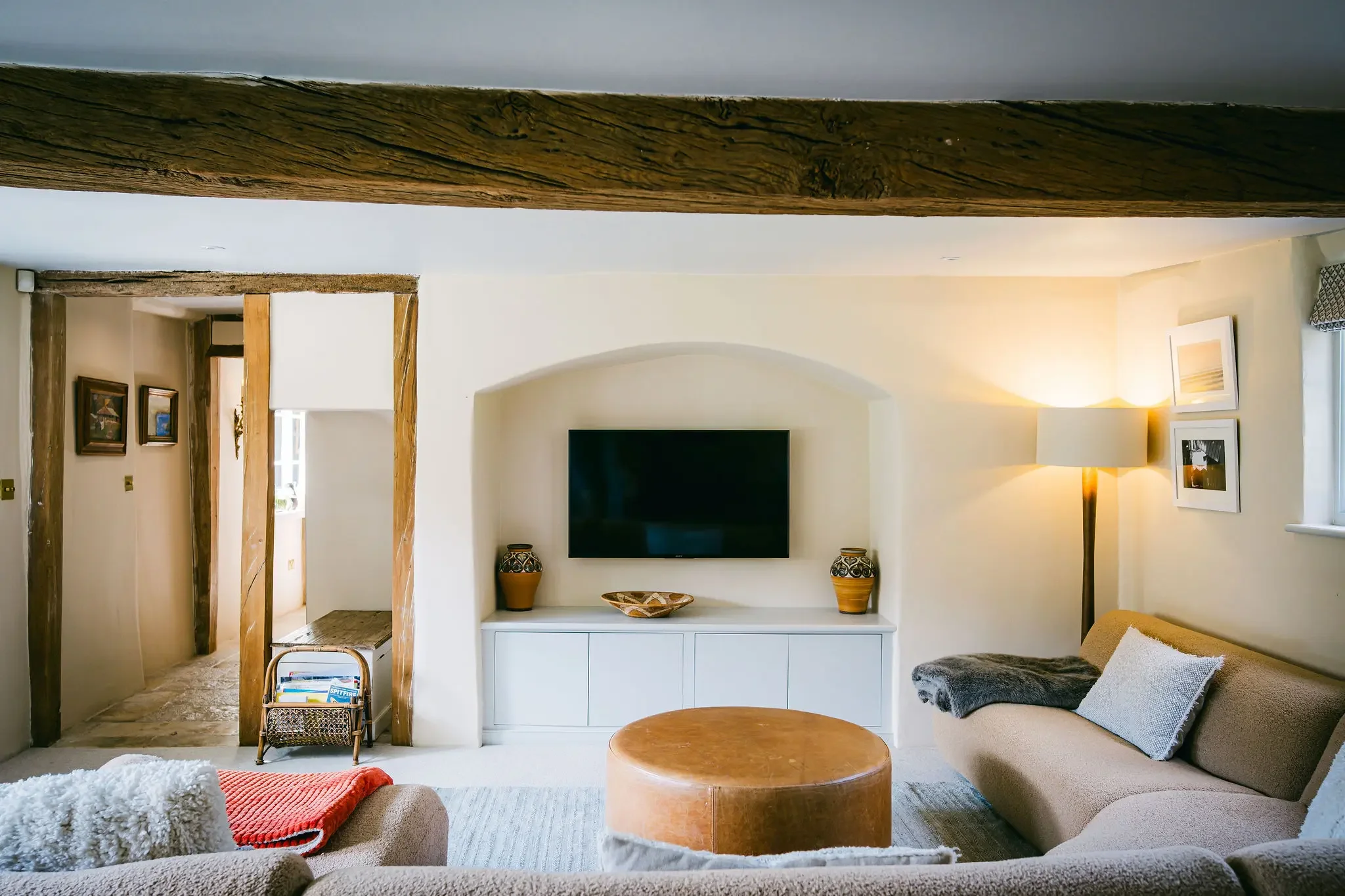 A cozy living room with a beige sofa, throw pillows, and a round wooden coffee table. A large flat-screen TV is mounted on a white wall, flanked by two decorative vases. There is a standing lamp and wall art, with wooden beams on the ceiling and entryway to another room.