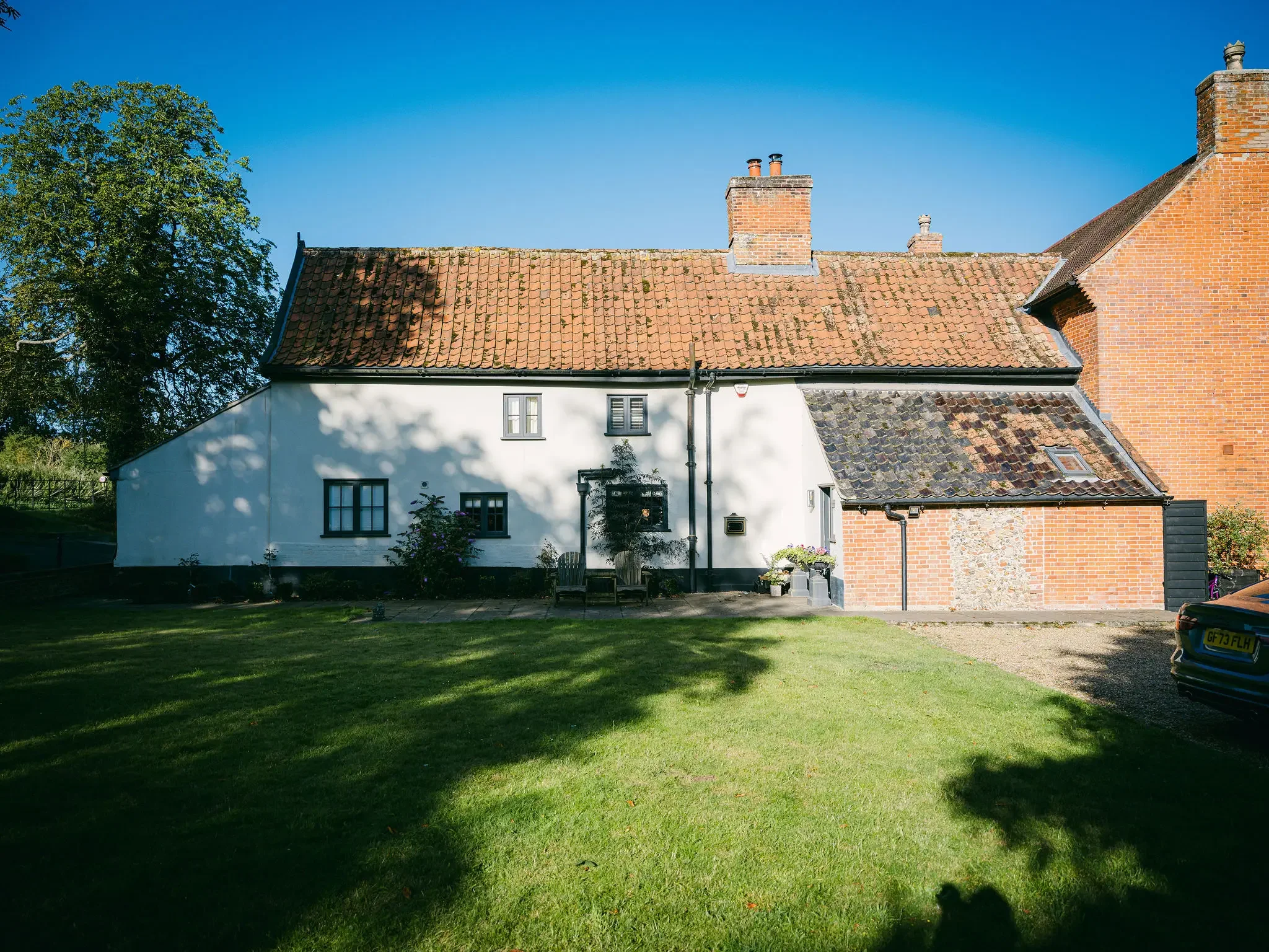A house with a white and red brick exterior, a tiled roof, and several chimneys, set against a clear blue sky and surrounded by a grassy yard with some trees and a parked car.