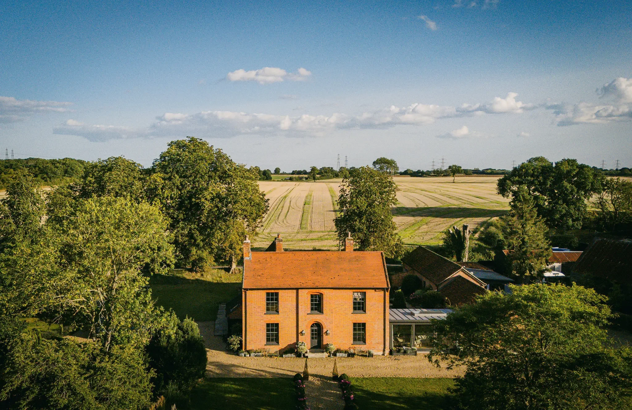 A large brick house with a red roof in a rural area, surrounded by trees and green lawns. In the background, there are fields and power lines under a blue sky with white clouds.
