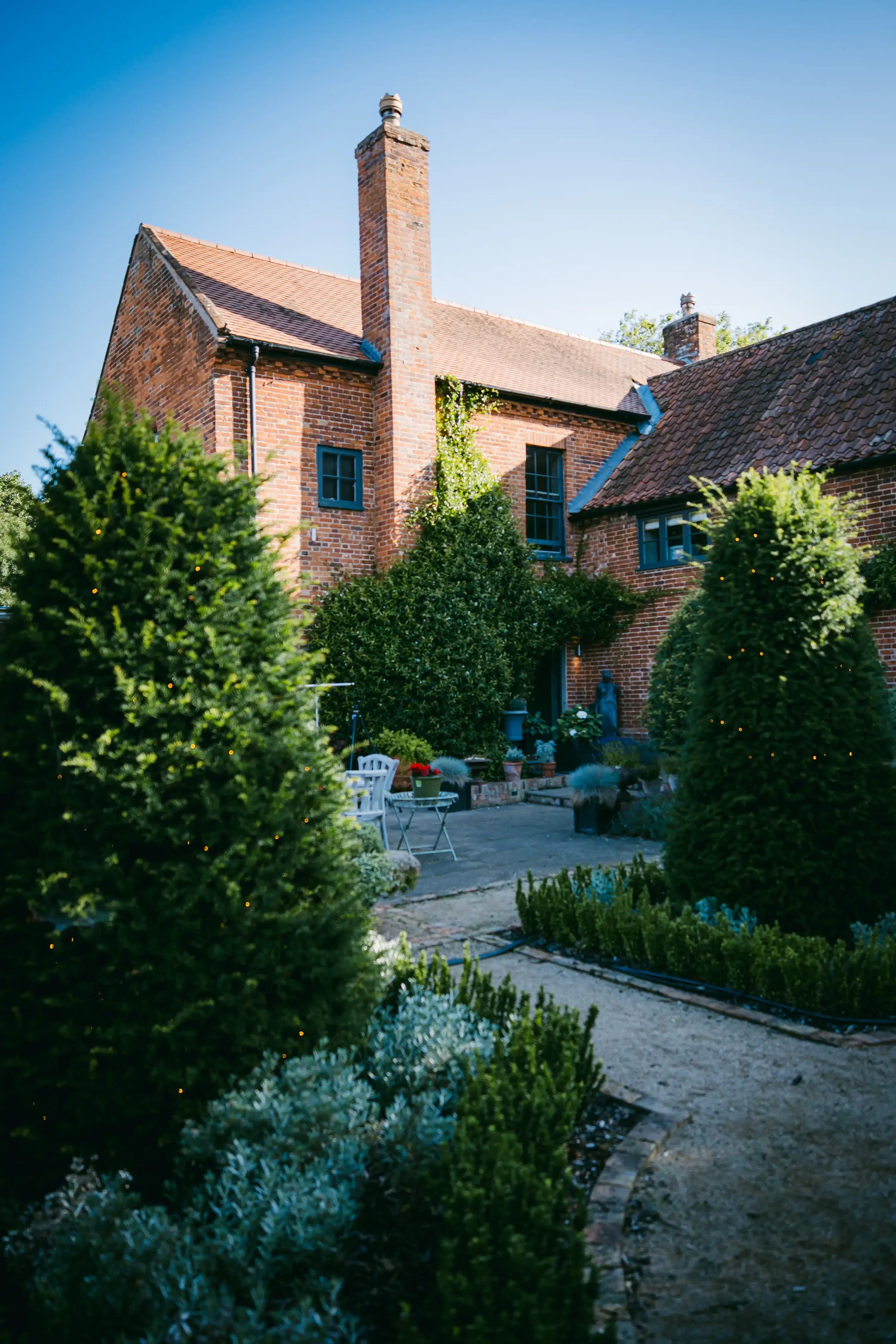 A brick house with a chimney, green garden, and outdoor seating area on a sunny day.
