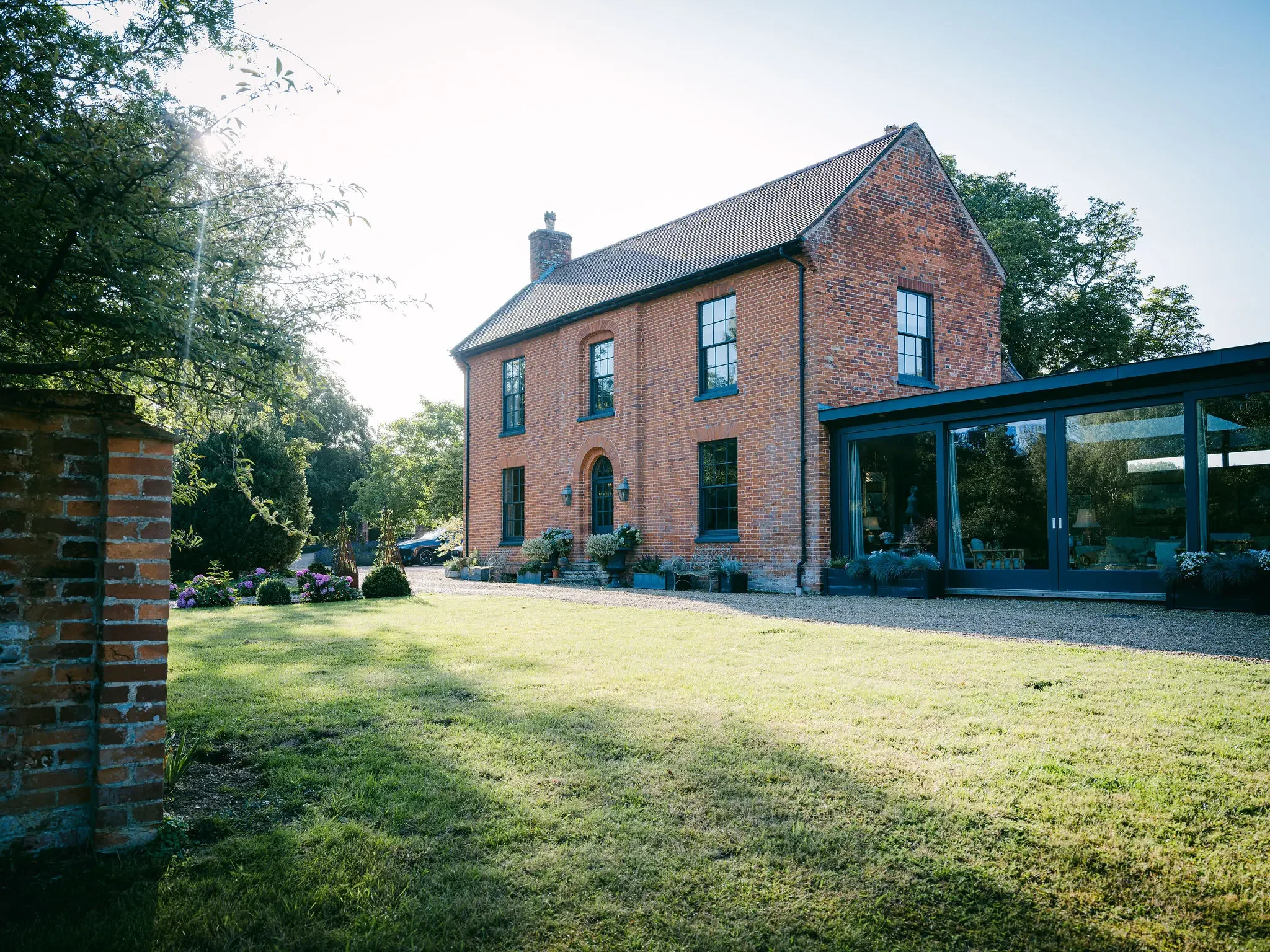 A brick house with tall windows and a sloped roof, surrounded by a green lawn and trees, with the sun shining from behind the trees.