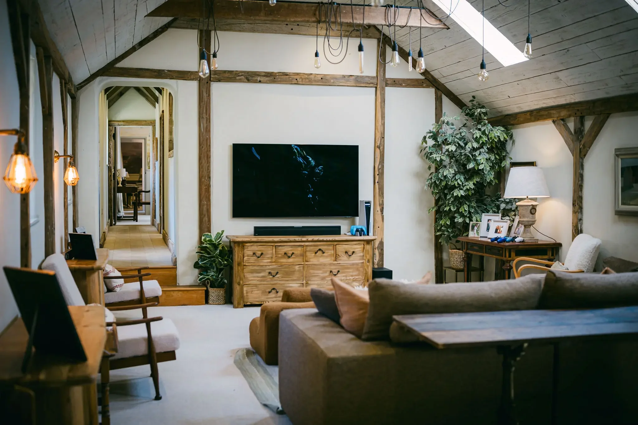 A living room with wooden beams, a large flat-screen TV, a wooden cabinet, a green potted plant, a table lamp, framed photos, and various chairs, with a hallway leading to other rooms.