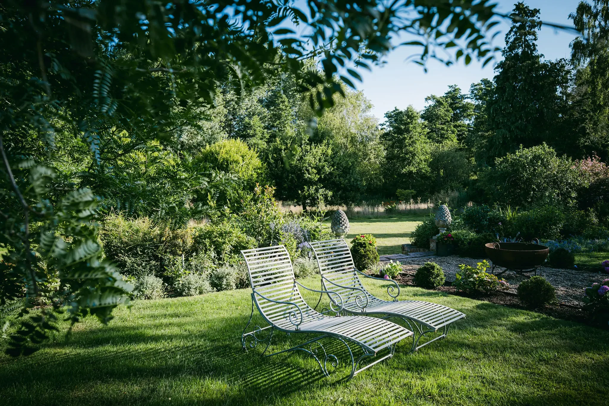 Green backyard with metal chairs, lush trees, and flowering plants, sunny sky visible in background.