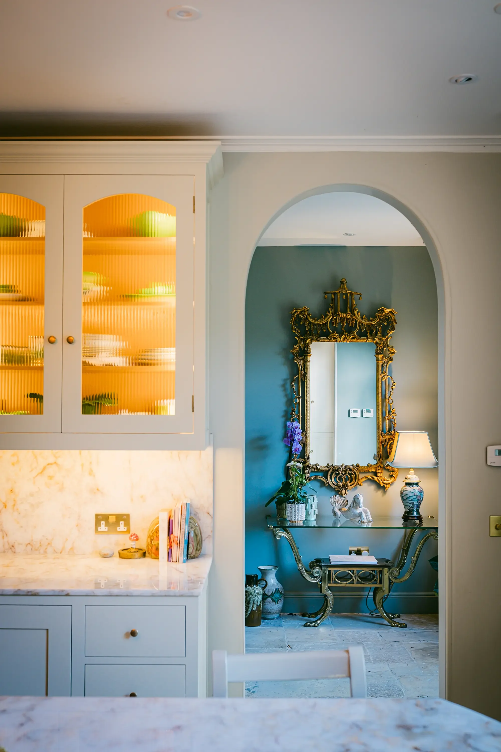 Interior view of a room with a marble countertop in the foreground, a built-in cabinet with glass doors on the left, and an ornate mirror and decorative table on the right through an arched doorway.
