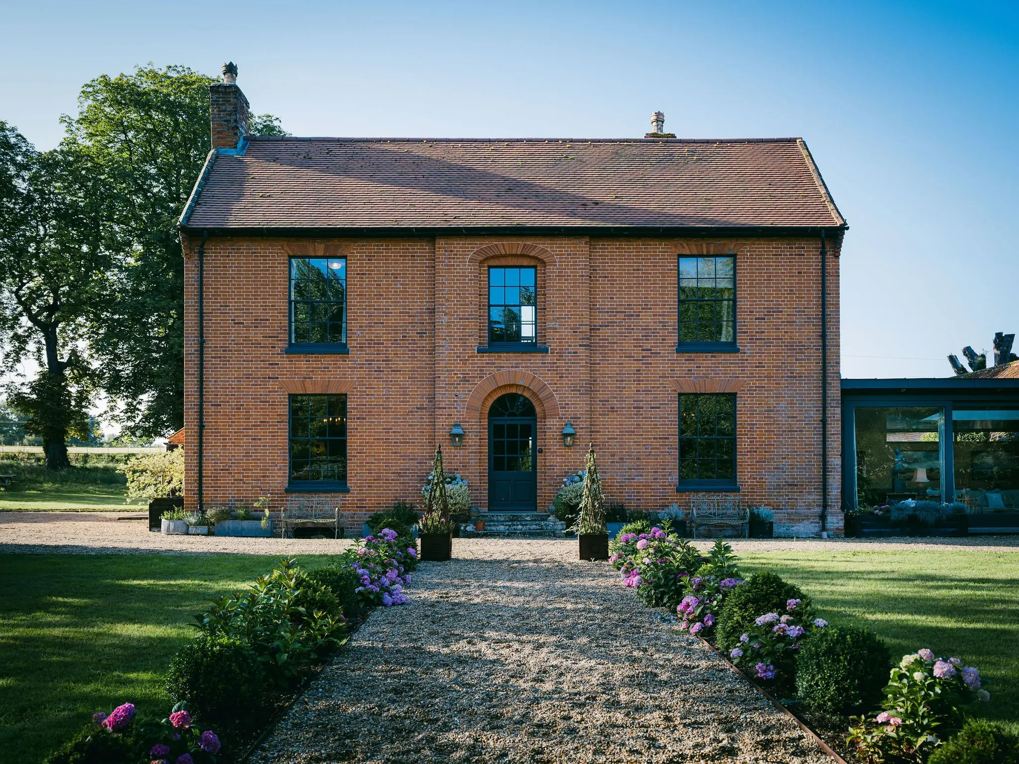 A brick manor-style house with large windows and a dark door, surrounded by a garden with pink and purple flowers, a gravel path, and green grass, under a clear blue sky.