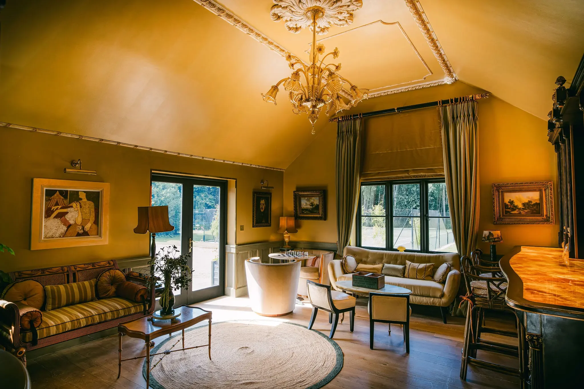 A cozy living room painted in warm yellow tones with elegant crown molding and decorative ceiling medallion. A gold chandelier hangs from the ceiling. The room features large windows with striped curtains, a cream-colored sofa, a round white armchair, and a striped sofa with pillows. There are framed paintings on the walls, a wooden coffee table, and various lamps, creating a warm and inviting atmosphere.