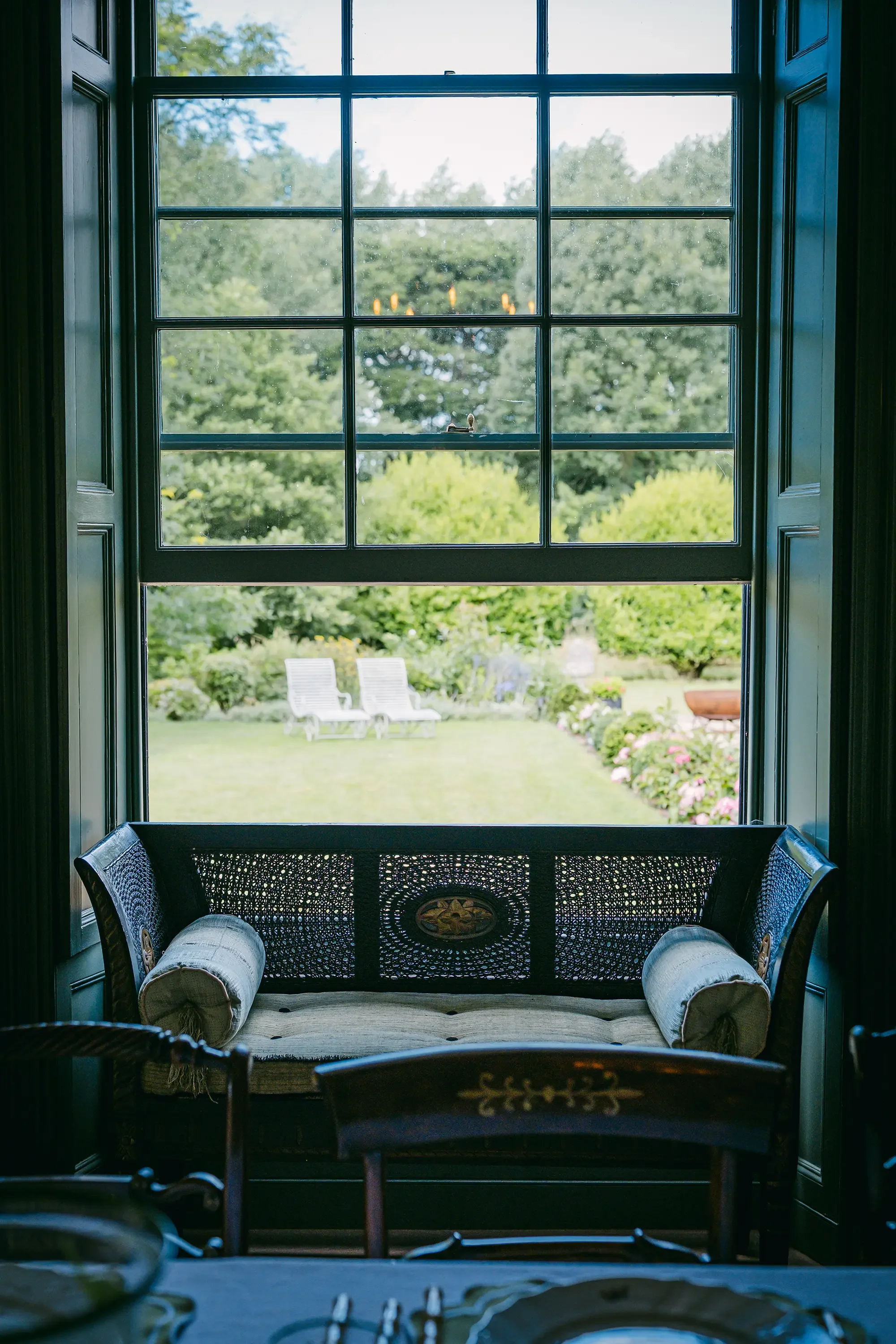 View through a large window showing a lush green garden with trees, bushes, and flower beds. Inside, in the foreground, there is a black woven daybed with decorative pillows and a cushioned armrest. The window has multiple panes, with a chandelier hanging outside visible through the glass.