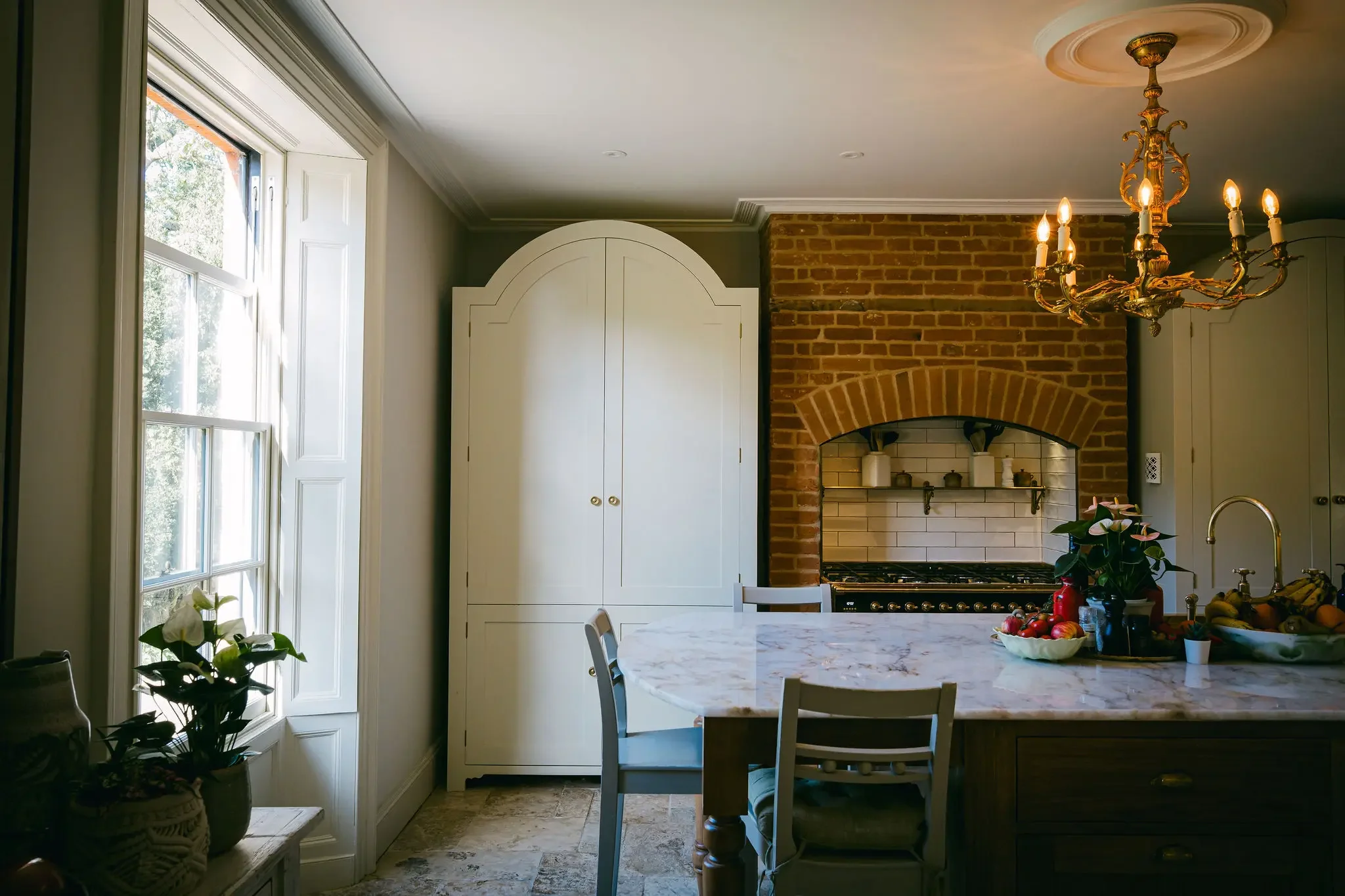 A cozy kitchen with a large window letting in natural light, a white cabinet, a brick stove hood, a marble-top dining table with chairs, a chandelier hanging from the ceiling, and a fruit bowl on the table.