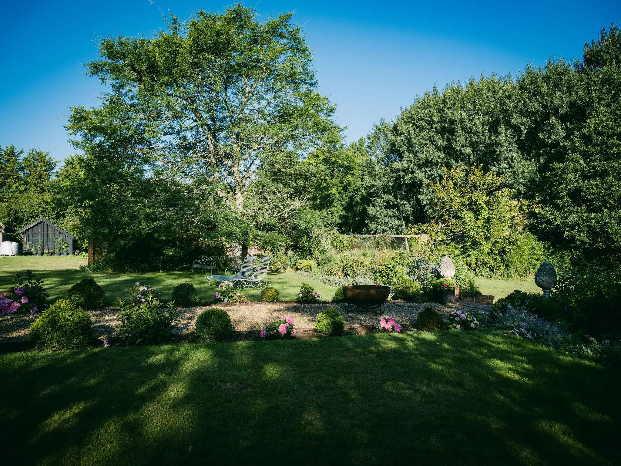 A lush green garden with various trees, colorful flowers, a small wooden shed, garden furniture, and decorative stone sculptures under a clear blue sky.