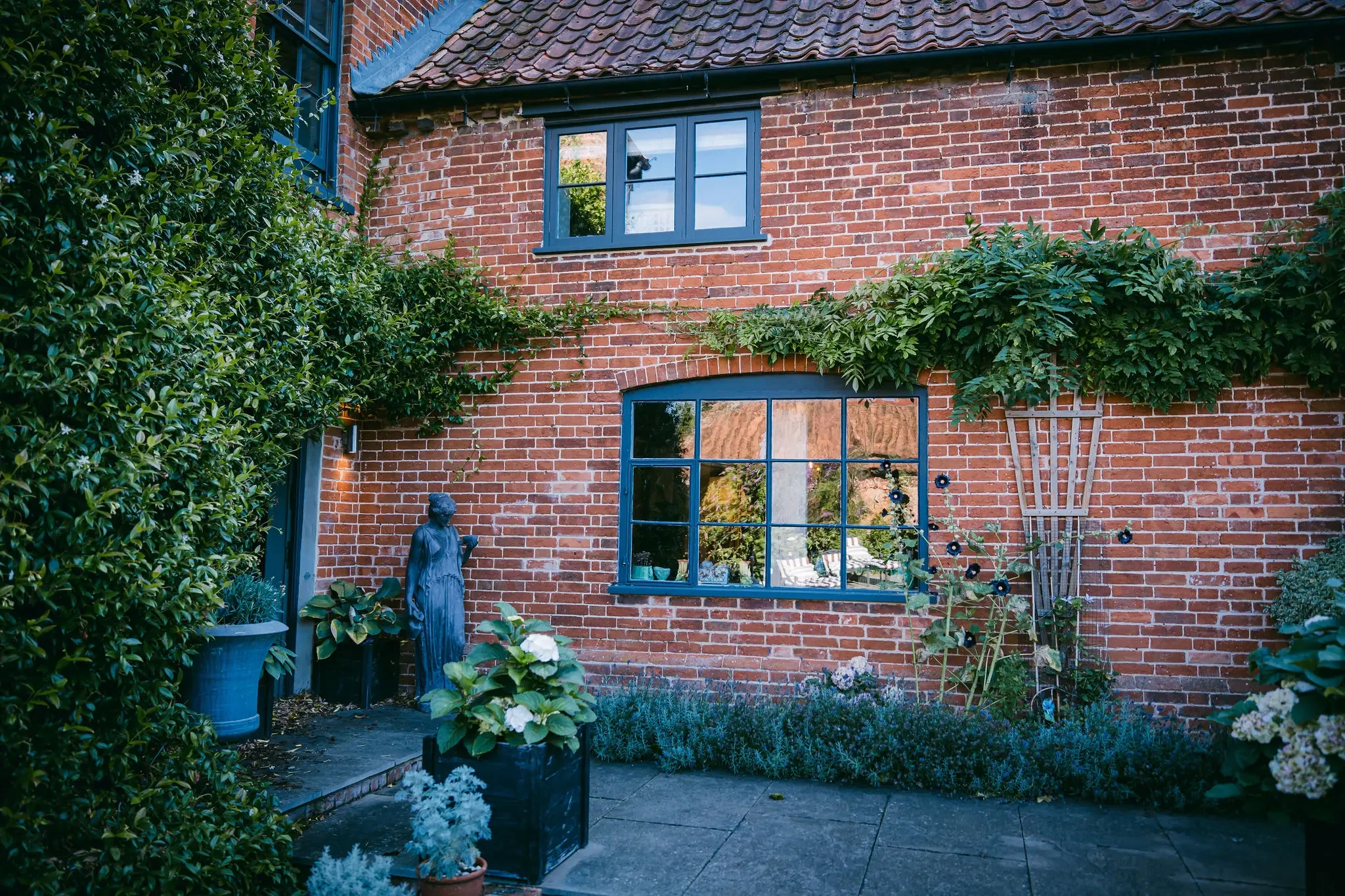 A brick house with large windows and a tiled roof. There is a garden with various plants and flowers, along with a statue of a woman. A trellis with climbing plants is attached to the wall.