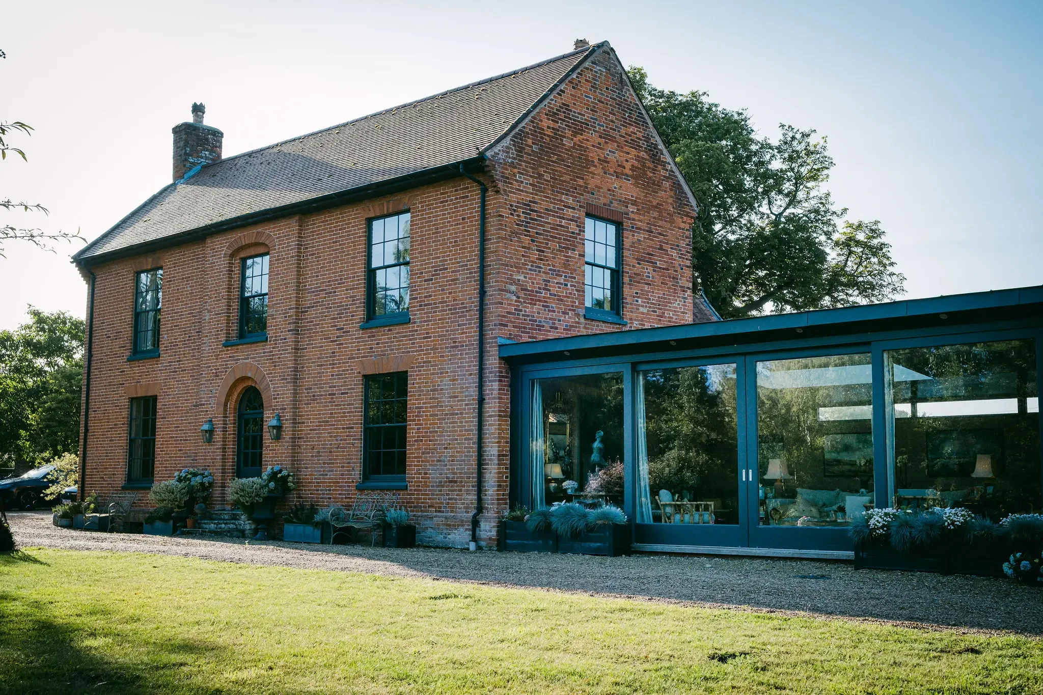 A large brick house with tall windows and a steep roof, adjacent to a modern glass extension, with a green lawn and trees in the background.