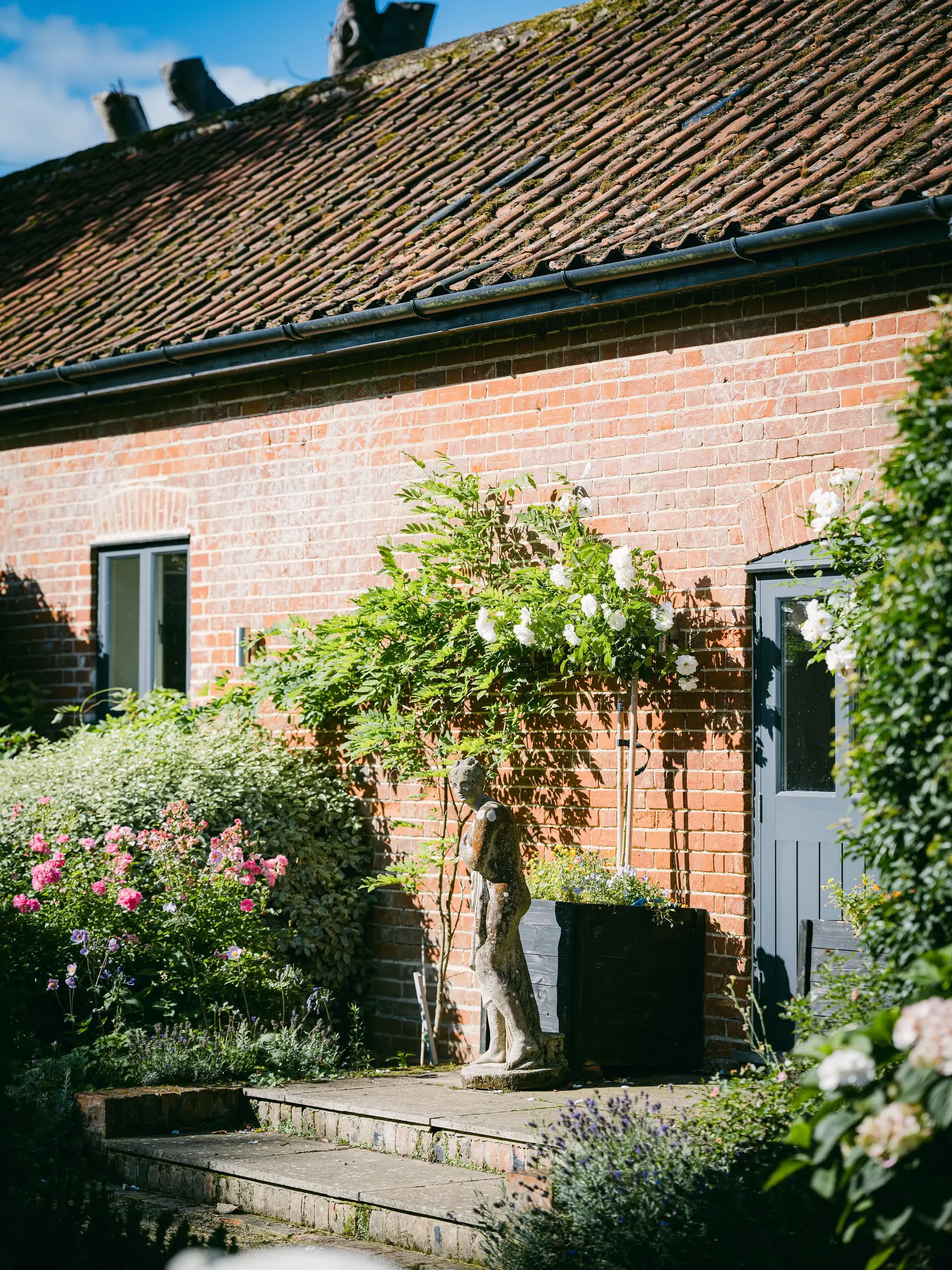A house with a brick exterior, dark window frames, and a blue door. A small garden with flowers and a sculpture of a person is in front of the house, with a mossy tile roof and a black gutter along the roof's edge.