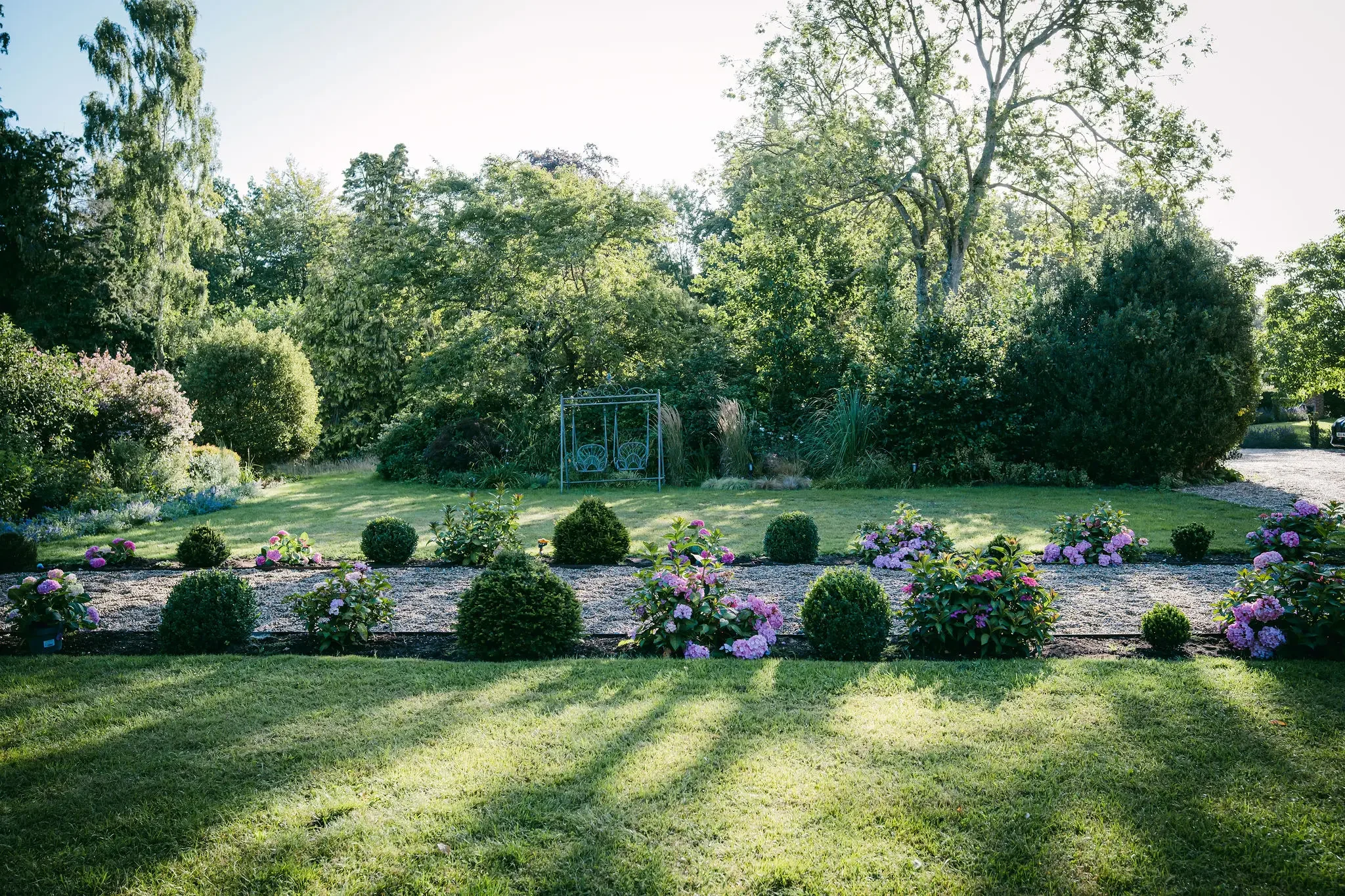A well-maintained garden with blooming pink flowers, small bushes, and neatly cut grass, surrounded by tall trees and a blue metal swing on a sunny day.