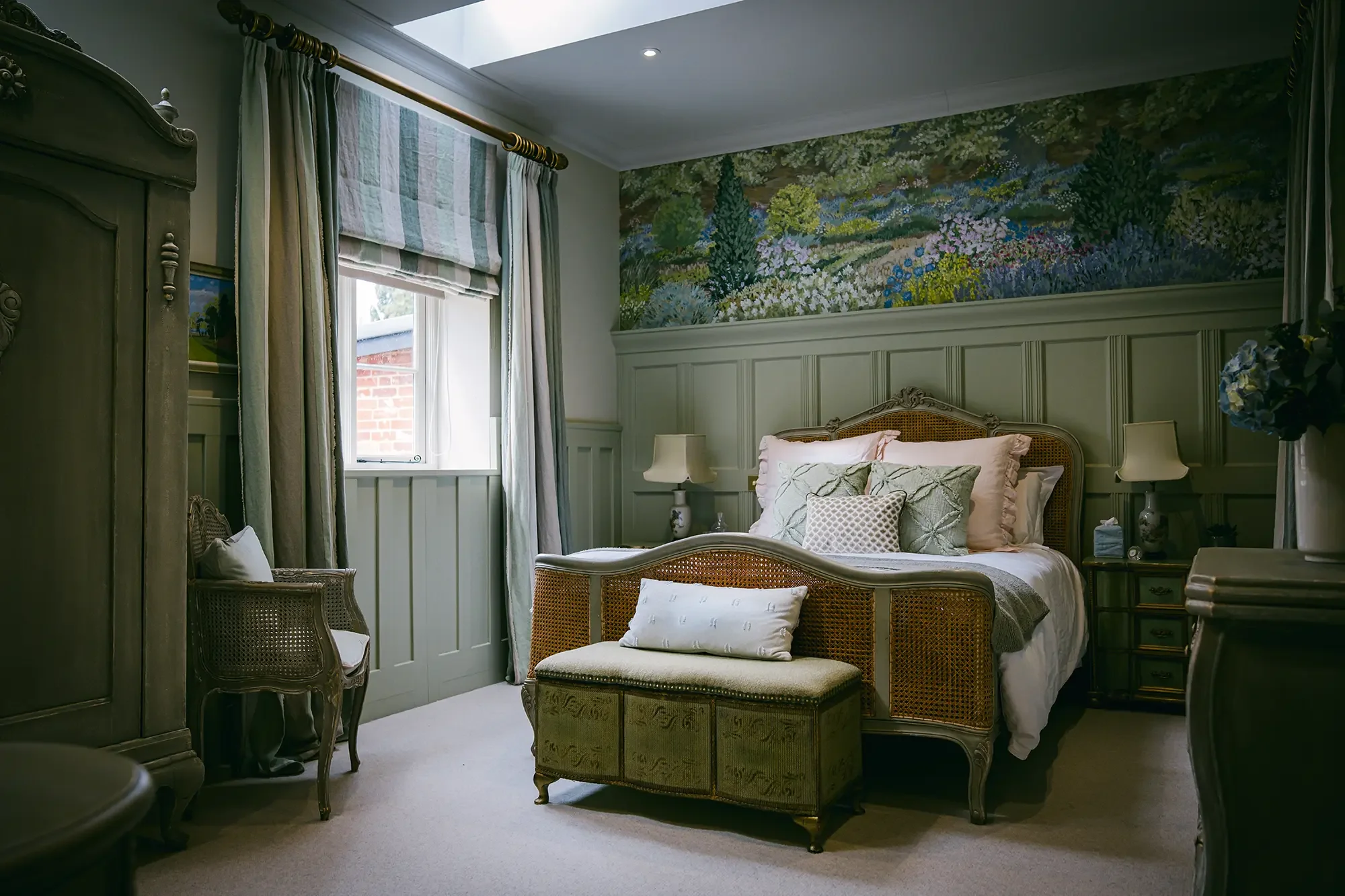 A cozy bedroom with a large bed that has a rattan headboard, adorned with pink and white pillows. There are bedside tables with lamps on either side of the bed. The room features wall paneling and a floral painting above the bed. A window with a striped Roman shade and curtains allows natural light into the room. An embroidered bench at the foot of the bed and decorative chairs are also visible.